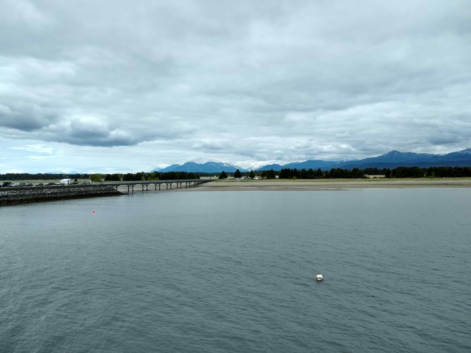 A long dock and jetty with some cars parked on it sticking out into the water. In the distance are some mountains and a few buildings of a small town.