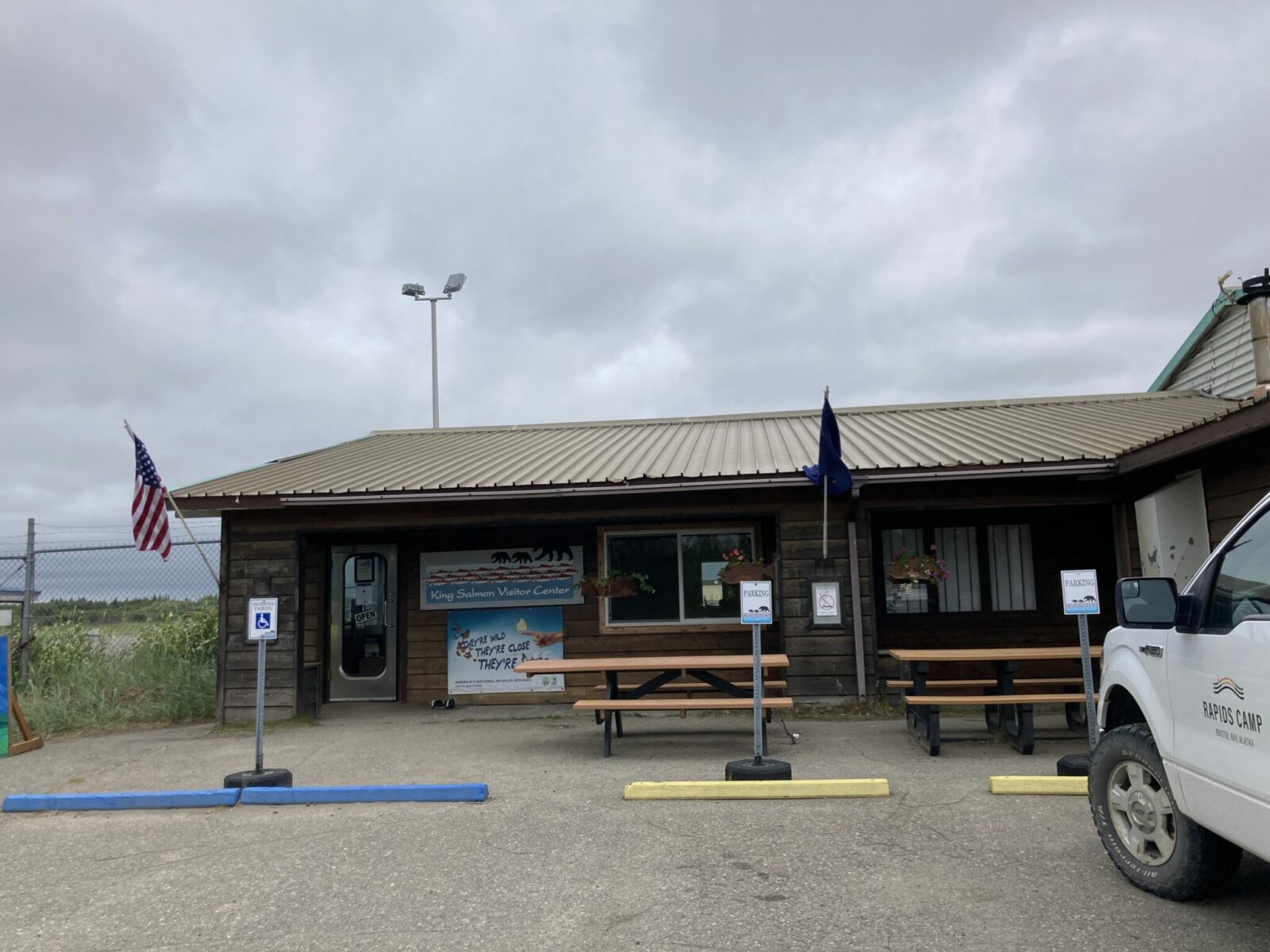 A small wooden building with an American and Alaska flag and a fence behind it. The signs reads King Salmon Visitor Center and has several bears drawn on it.