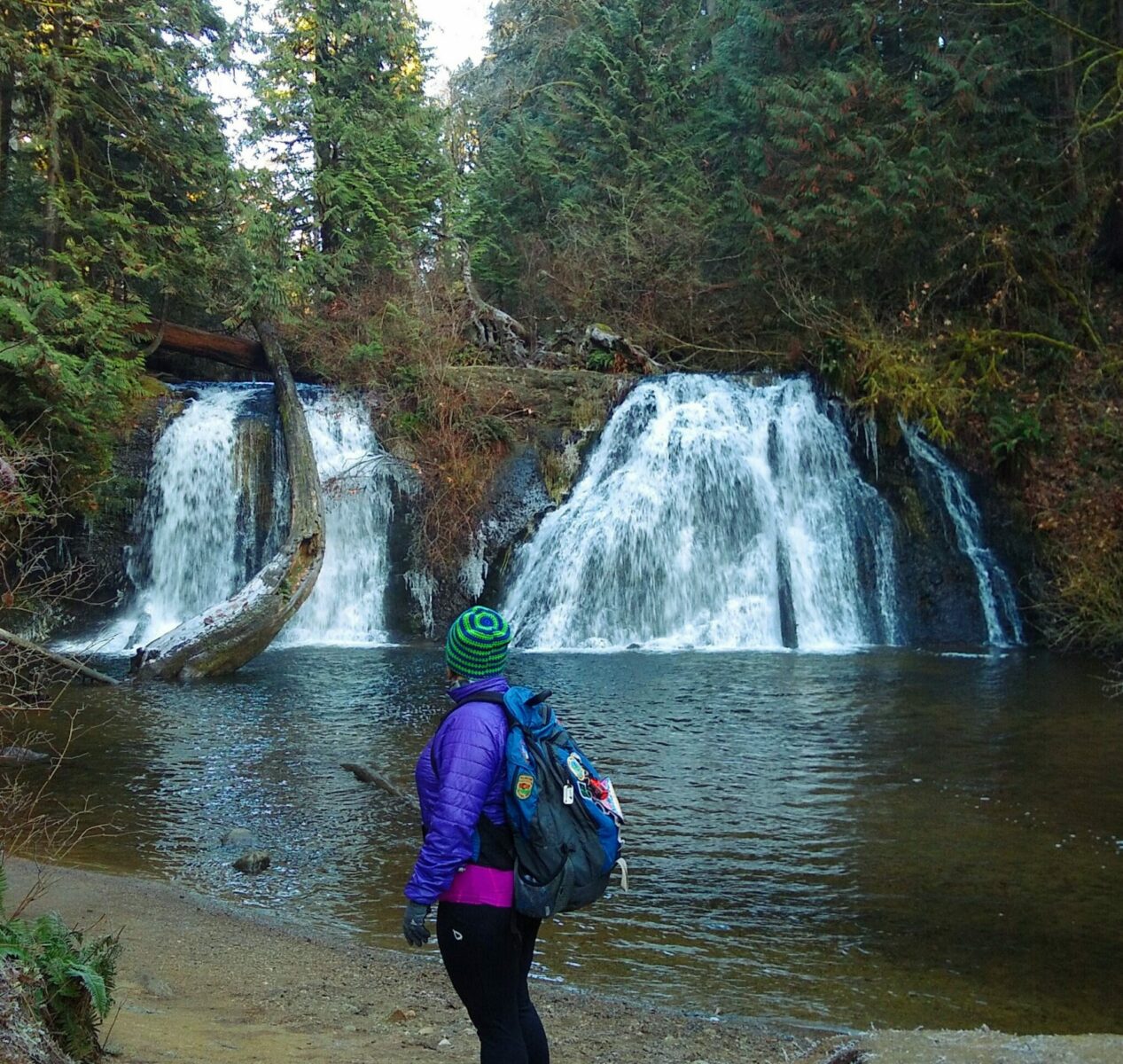 A hiker in a winter hat and coat with gloves and a backpack standing on a gravel beach looking across a small river to a waterfall in the forest