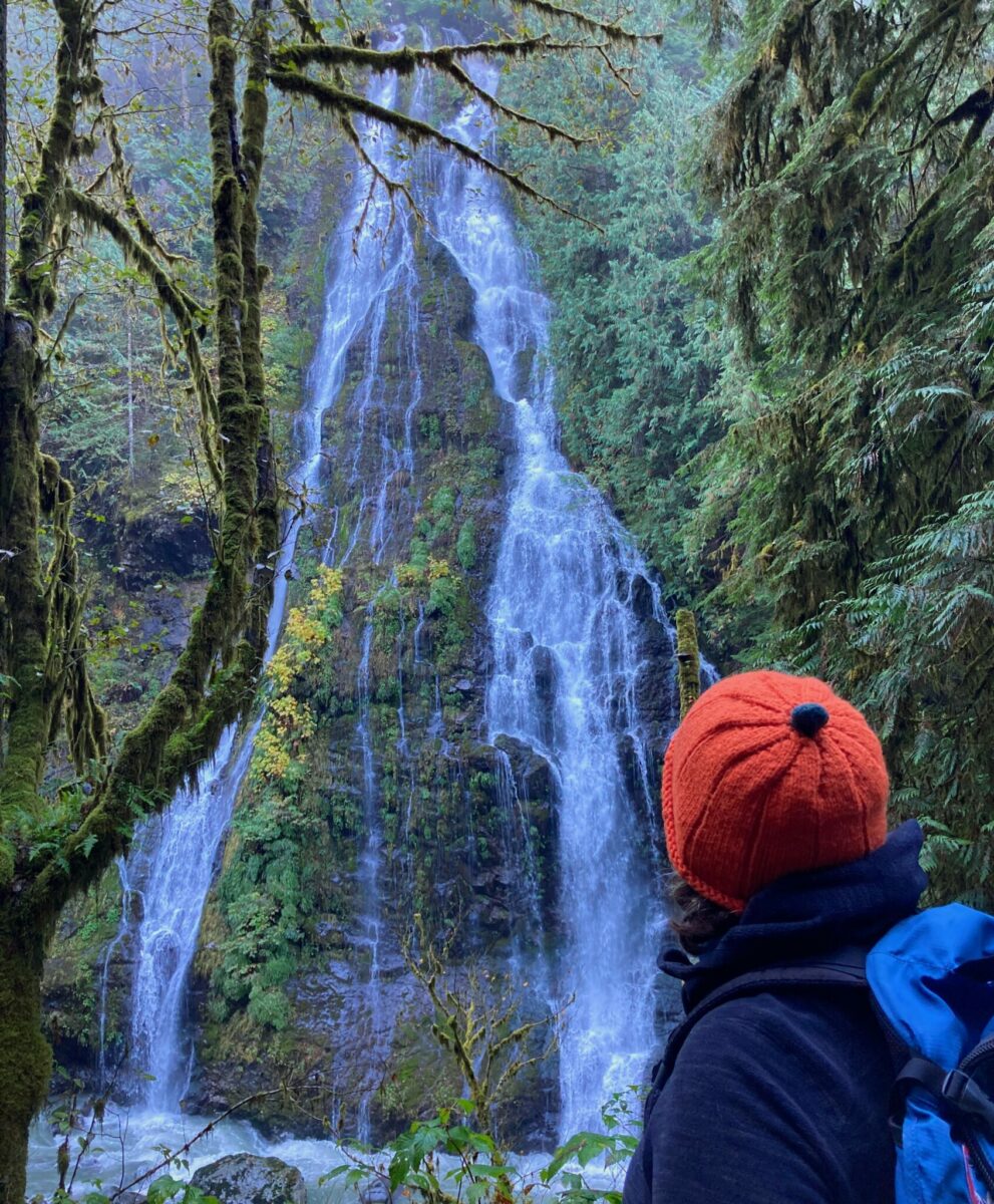 A hiker in an orange hat looking across a creek to a high waterfall coming down over moss covered rocks in the forest