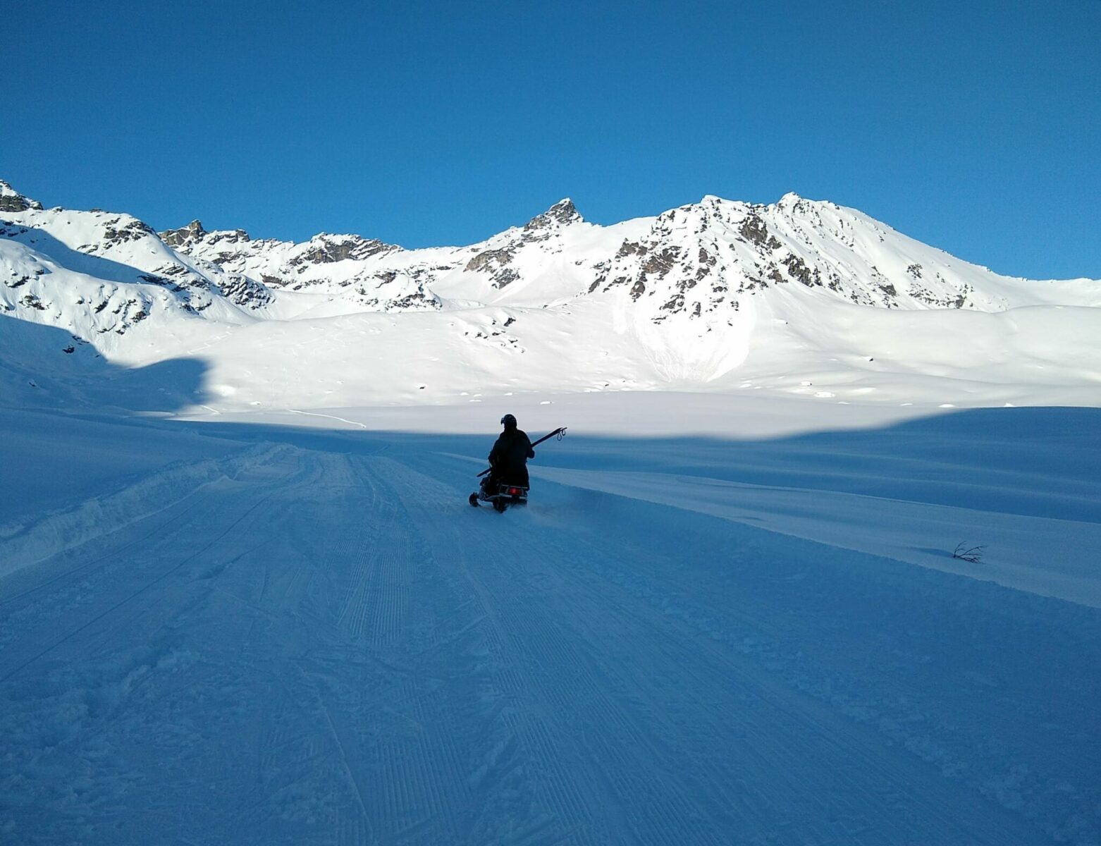 A snowmachine with a rider and a pair of cross country skis on a groomed ski trail with snow covered mountains in the background.