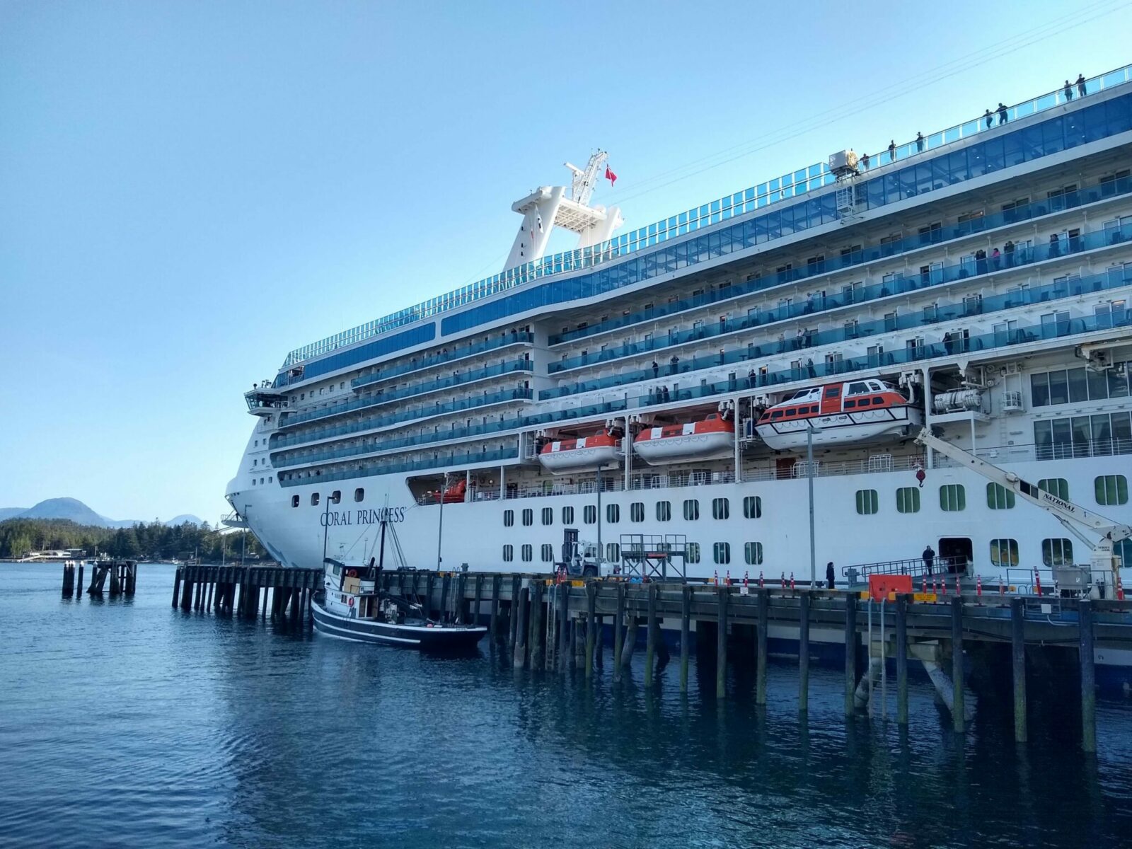 a cruise ship at a dock with a couple of smaller boats docked near by. You can see some forest and a few hills in the background.