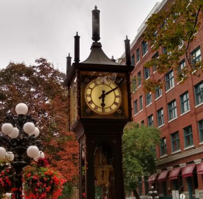 A historic steam clock on a street in front of a historic building and colorful trees.