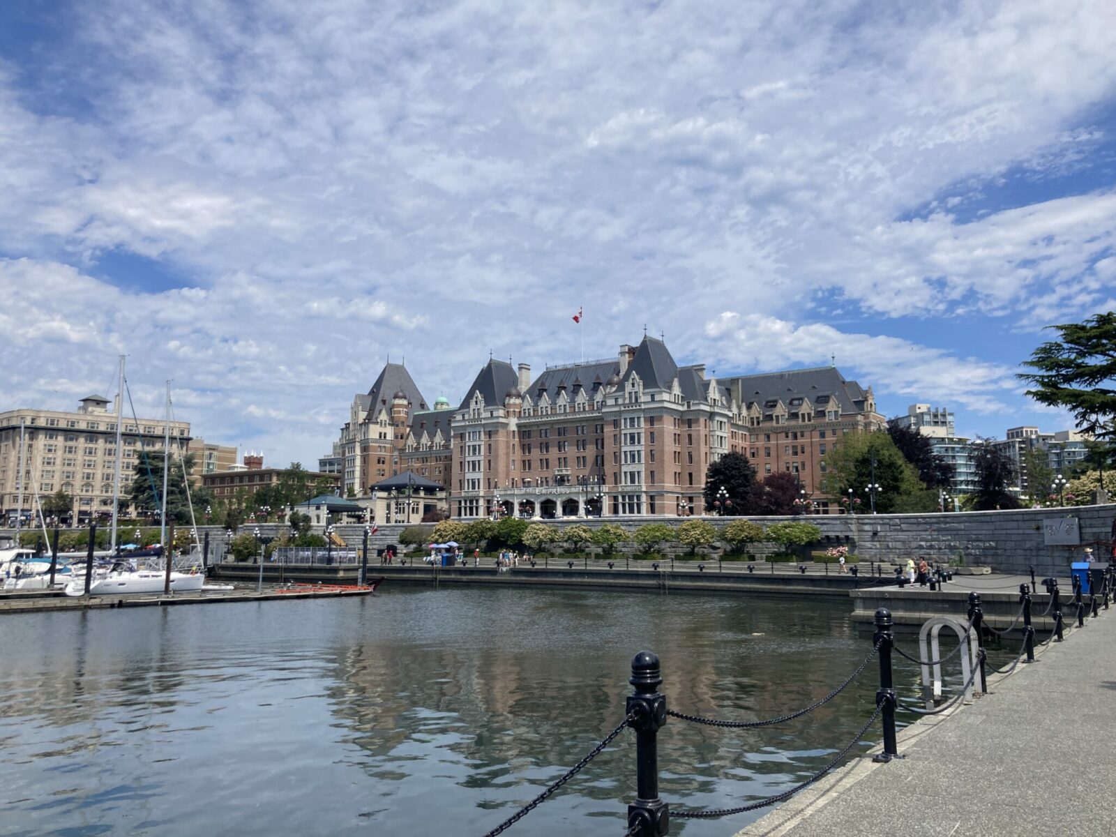 A historic hotel with brick and a metal roof. It is sitting along the edge of a harbor with a walkway between the hotel and boats