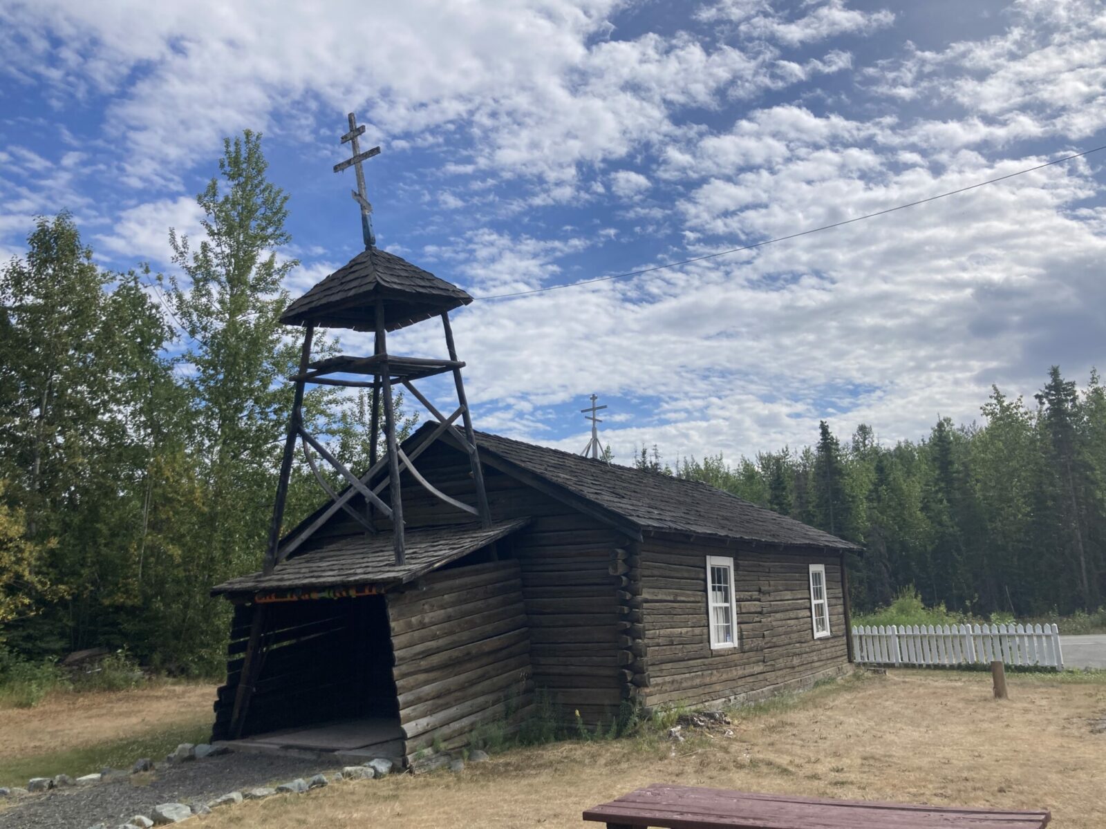 A historic log Russian orthodox church from the late 19th century. It has a wooden bell tower with a russian orthodox cross on top. it is surrounded by a white fence and a forest.