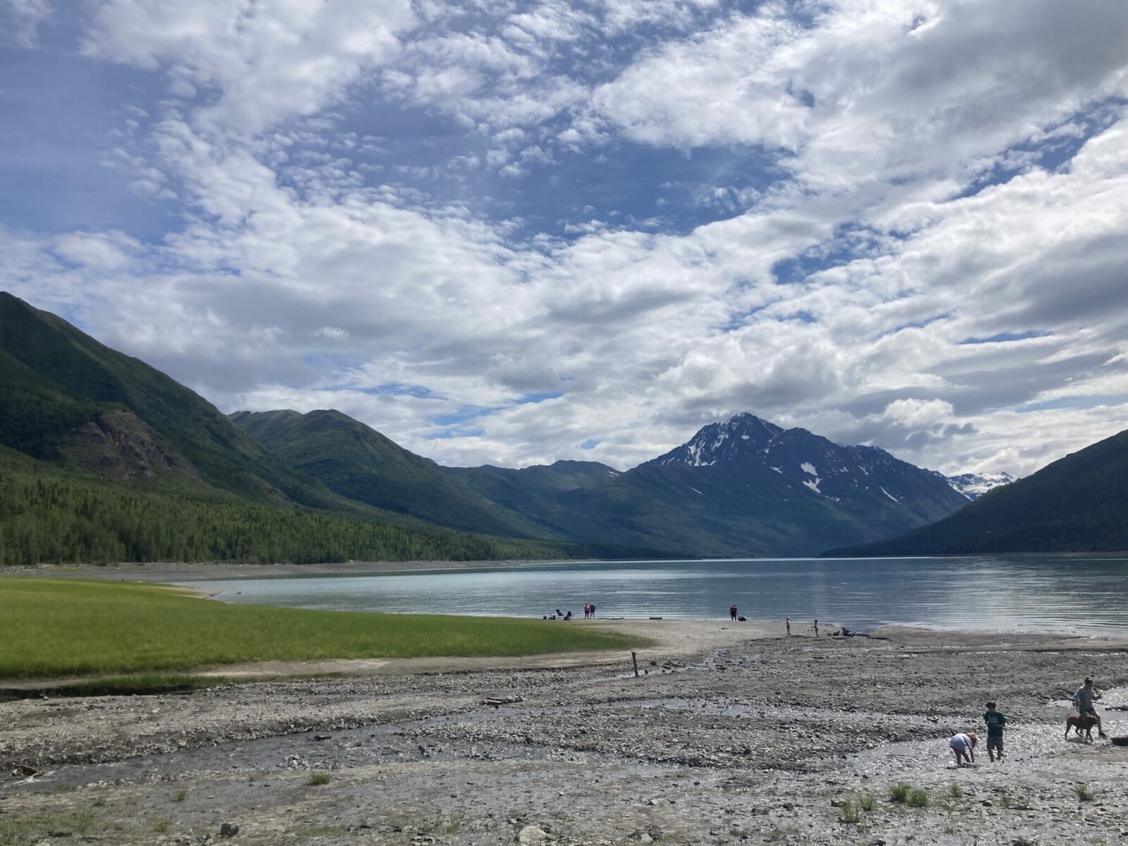 A gravel beach with green grass at the edge of Eklutna lake. In the distance are forested mountains and a bit of lingering snow on higher mountains on a partly cloudy day.