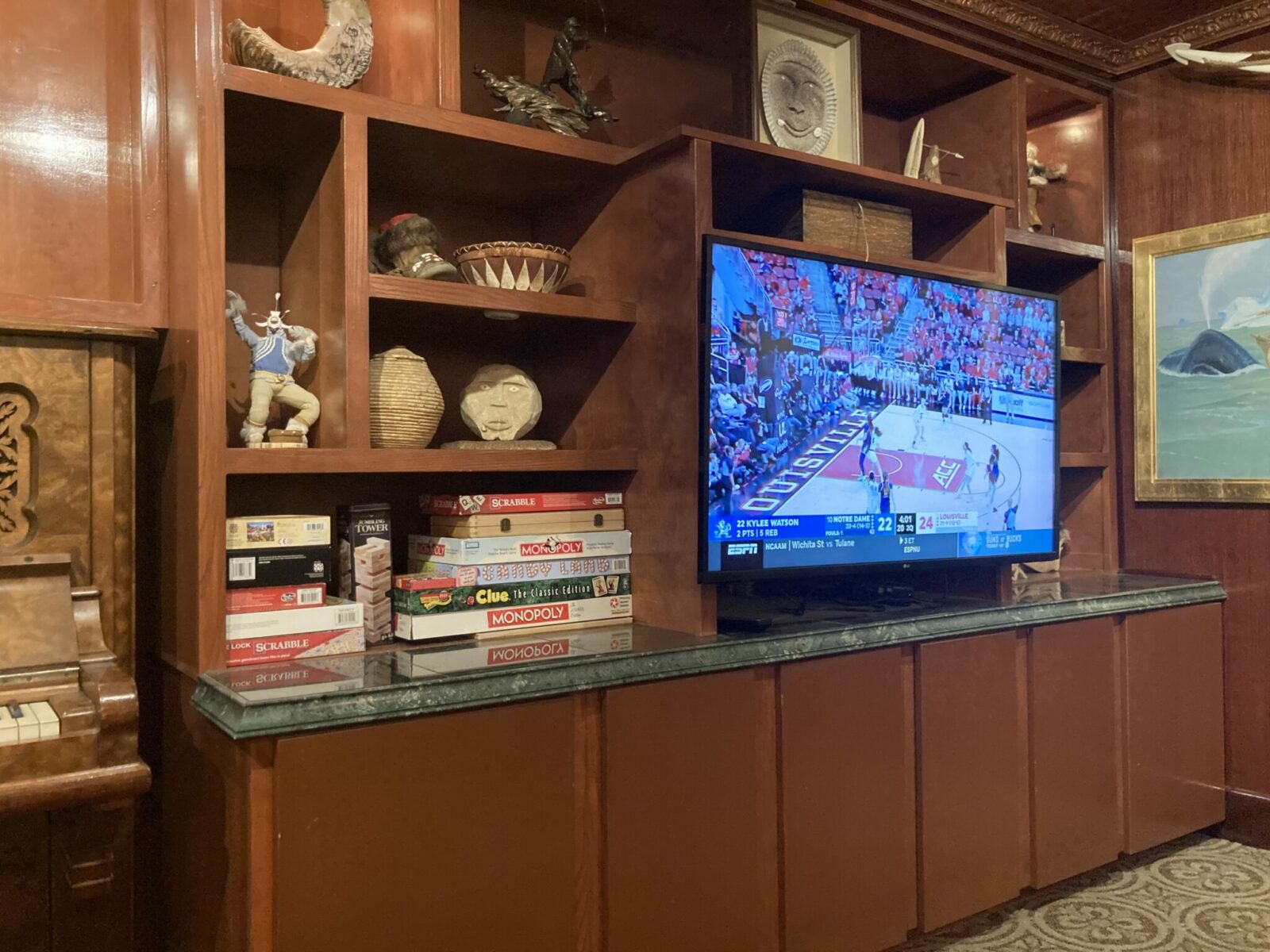 A wooden bookcase with Alaskan art as well as board games and a tv showing basketball