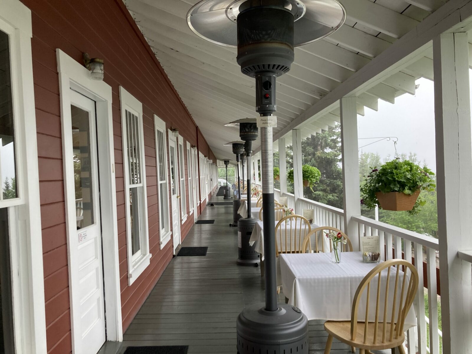 A wide covered wooden deck next to a red and white building. There are trees around the deck and several heaters and tables with flowers and white tableclothes.