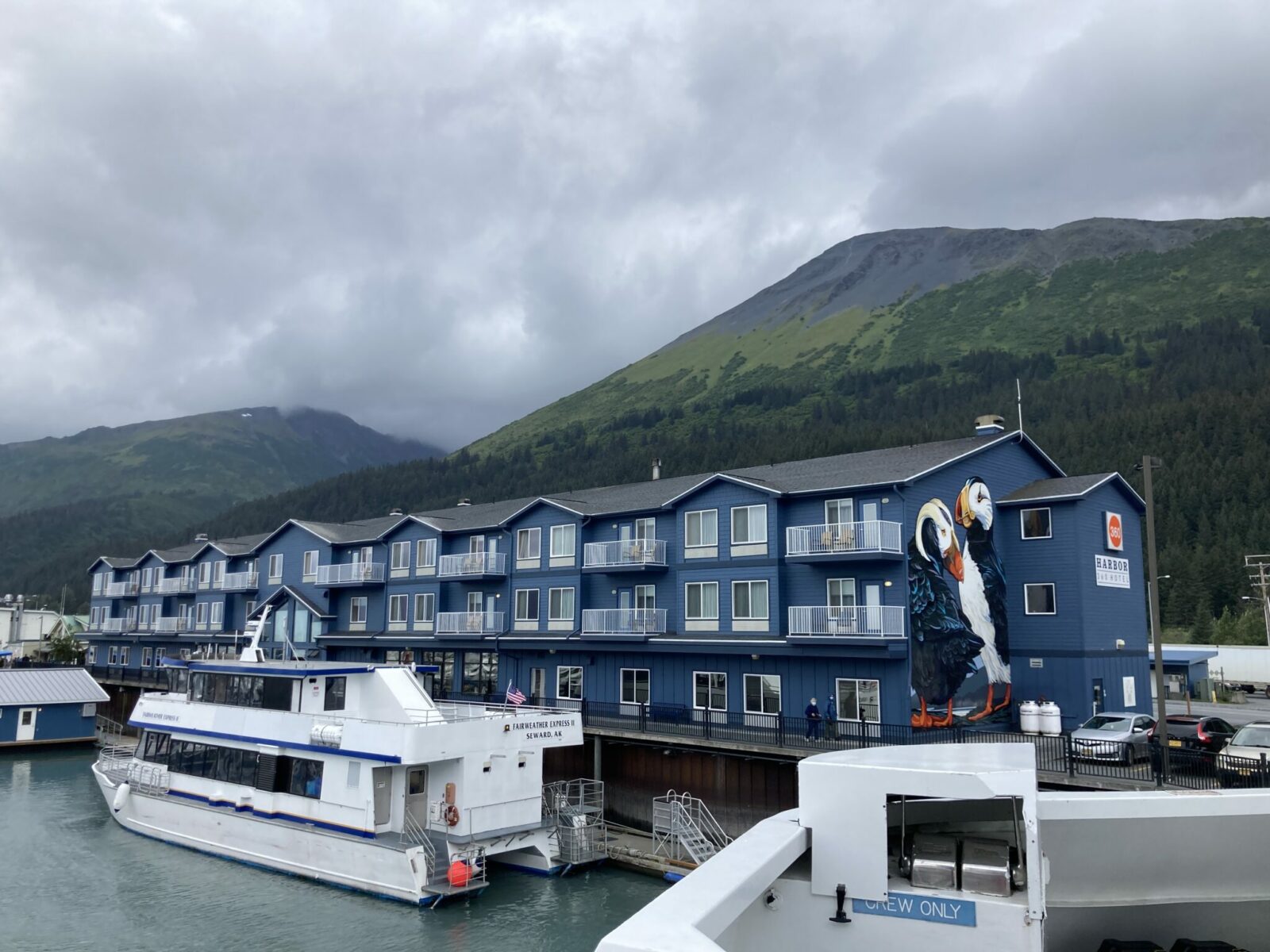 A blue hotel building with balconies on the waterfront with boats in front. In the background is are forested hillsides and mountains on a cloudy day. 