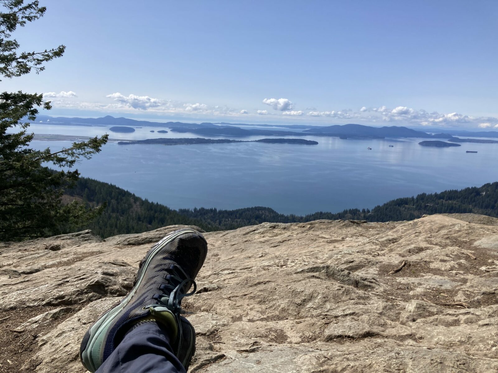 A hikers feet on the rocks above a big view down to the forest below as well as out to the water, forested islands and ships at sea.