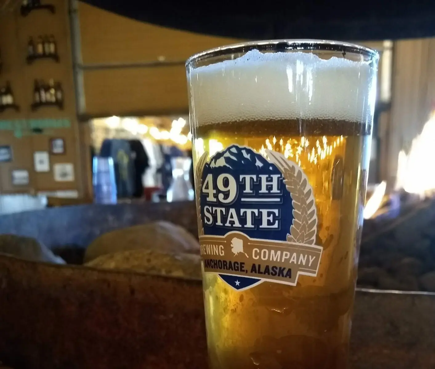 A beer in a clear glass on a table. The pint glass says 49th State brewing company Anchorage Alaska