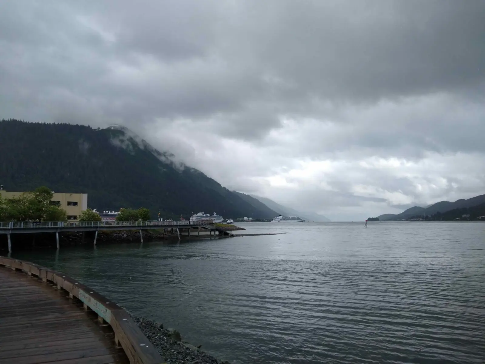 A cloudy and rainy day in Juneau Alaska. The view is of a wooden boardwalk along the shoreline with a few buildings and cruise ships in the background. The surrounding mountains are hidden by clouds and fog.