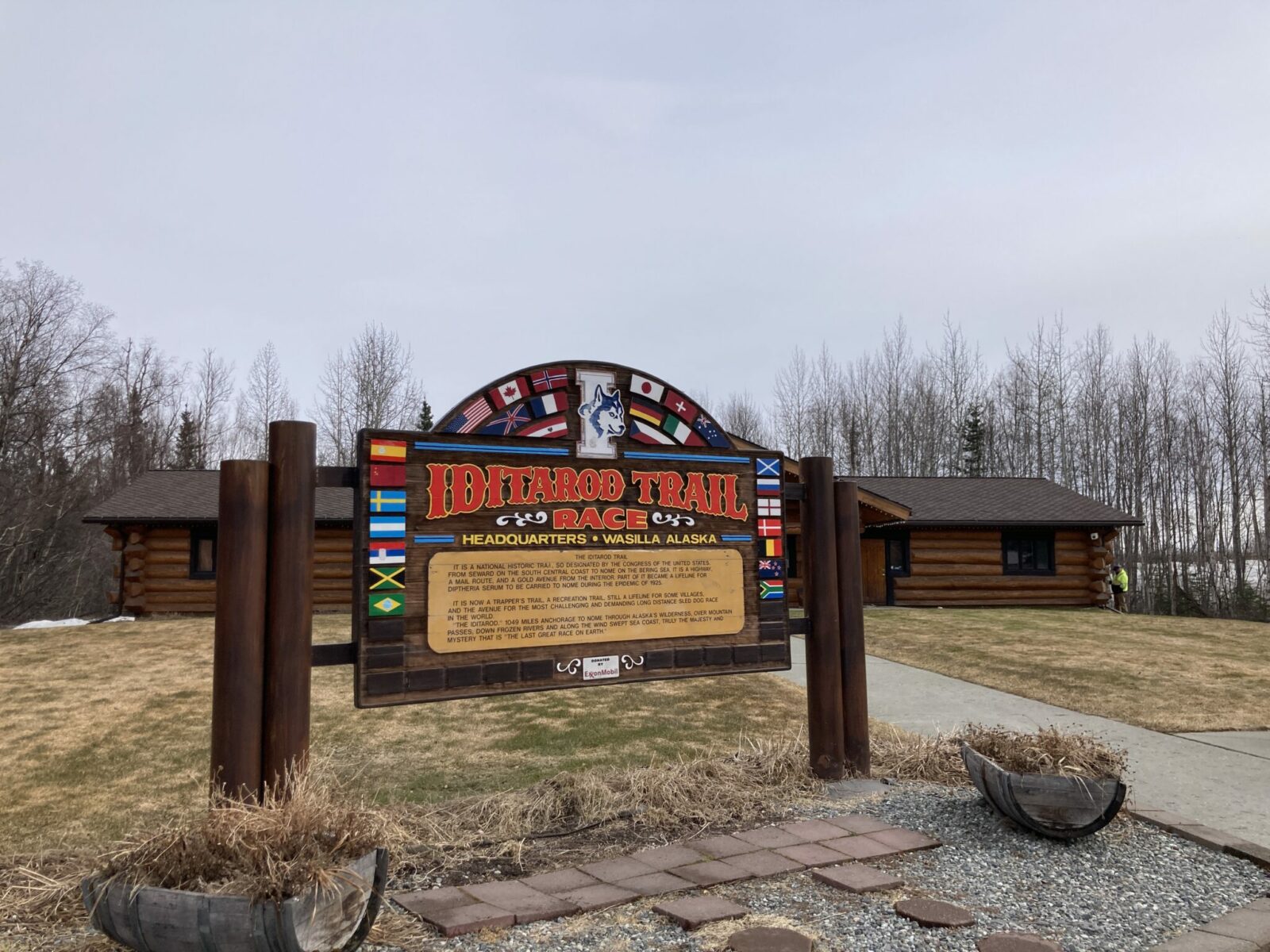 A log building surrounded by lawn with a sign in front with flags from around the world, the Iditarod logo and says Iditarod Trail race headquarters with information about the Iditarod.