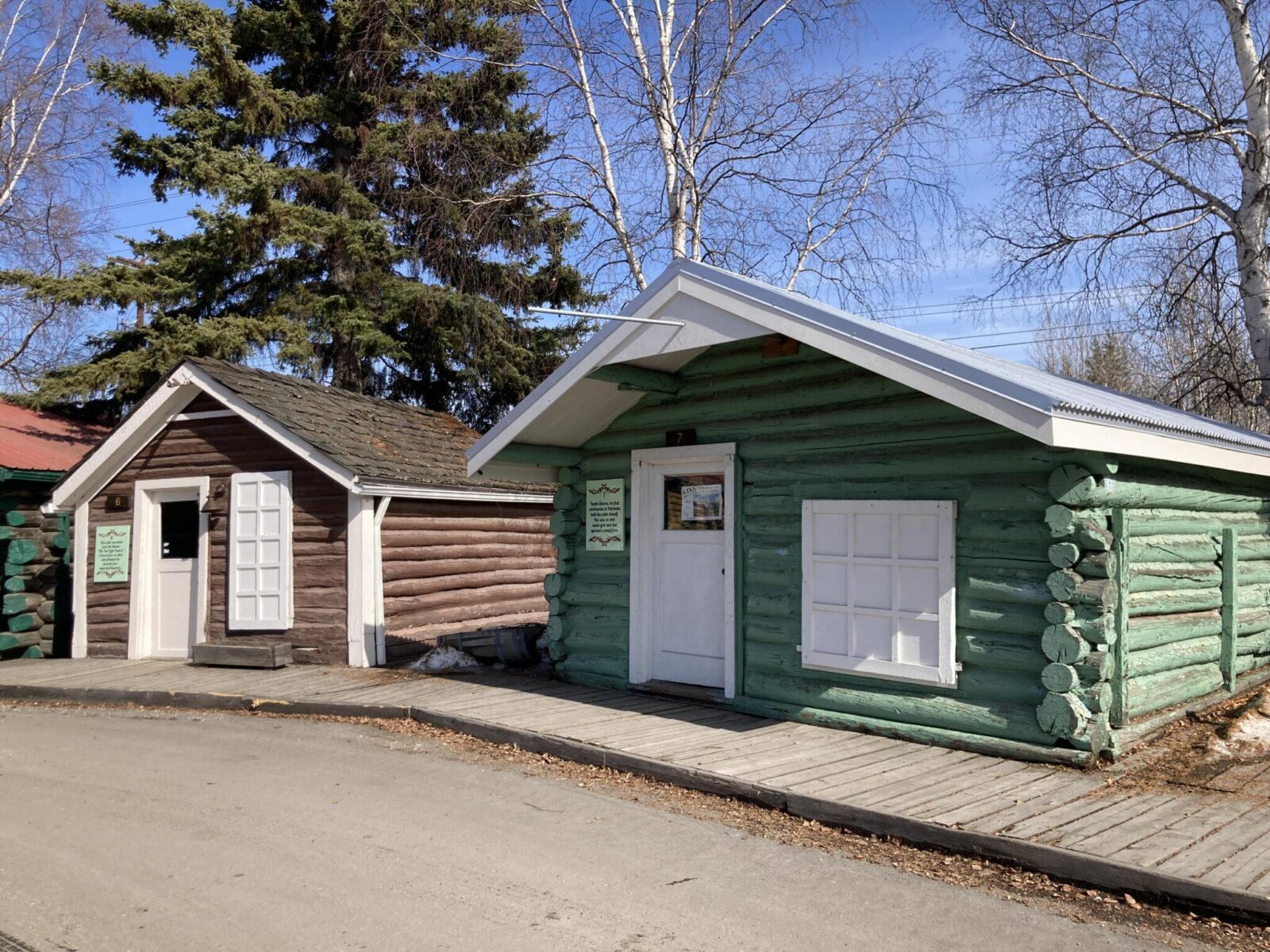 Two small, wooden cabins from the early 20th century in Pioneer Park in Fairbanks. One is brown and one is green and a wooden sidewalk connects them.