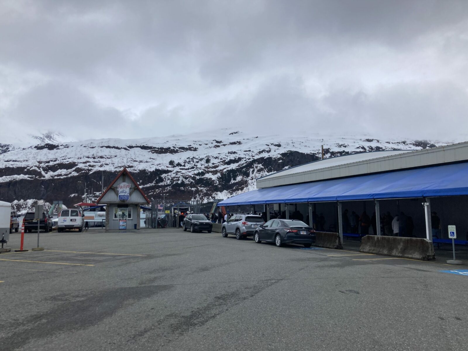 A small building with check in in a parking lot next to a covered waiting area. There are snowy hillsides in the distance