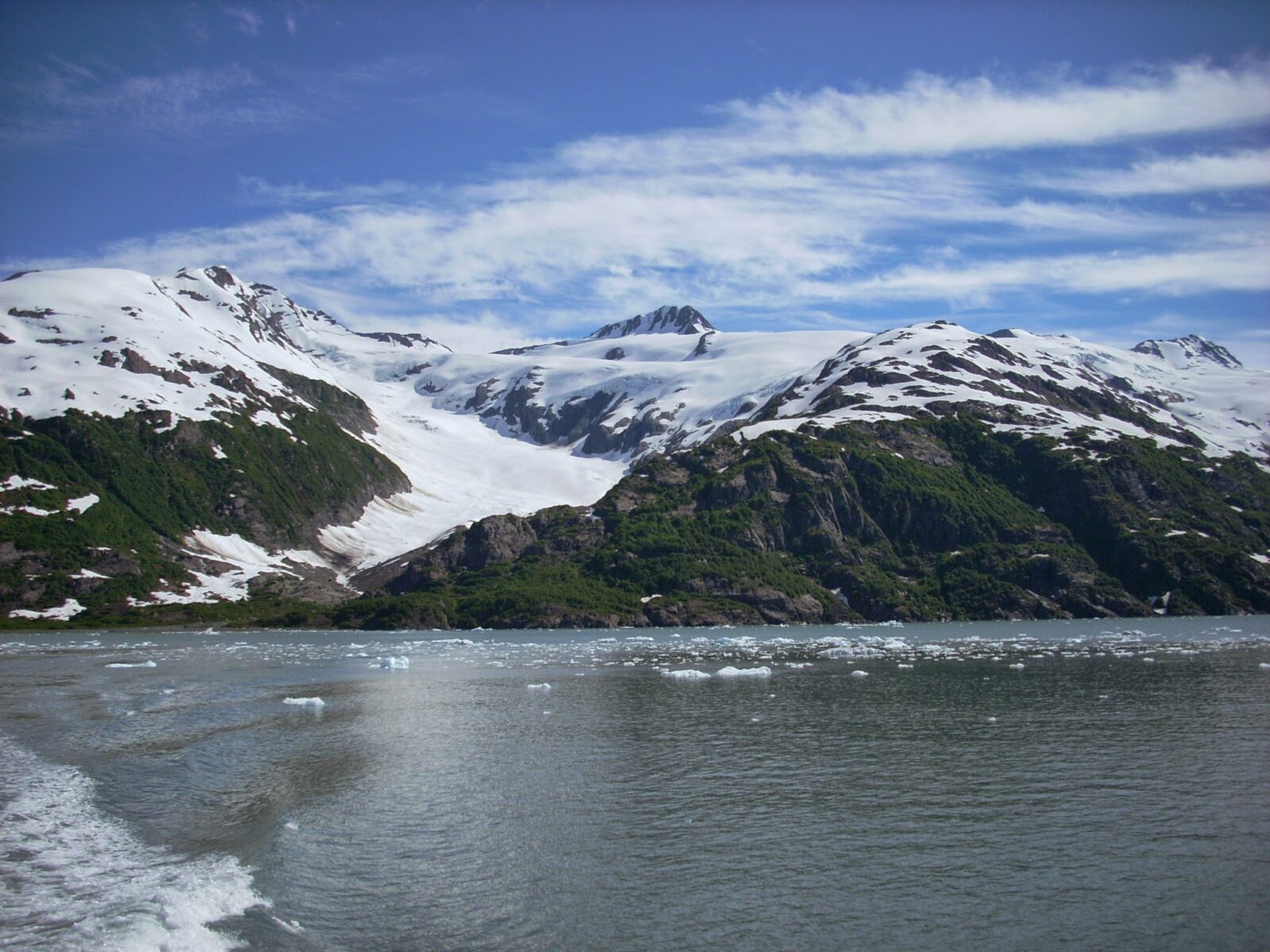 Icebergs in the water and a green hillside with several glaciers and mountains above it on a sunny day