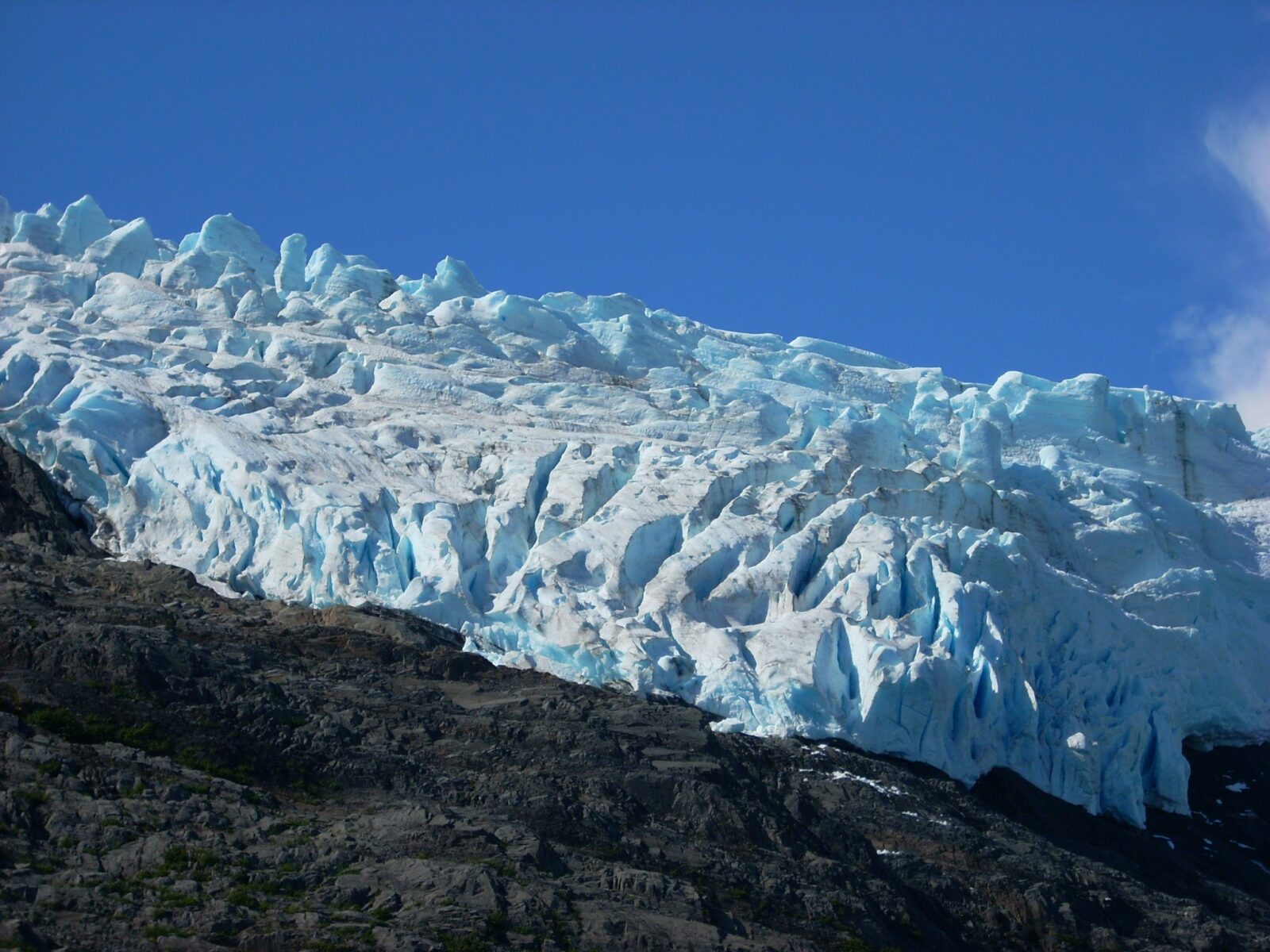 The edge of a hanging glacier seen from below. The ice is jagged and a combination of white and blue against a blue sky