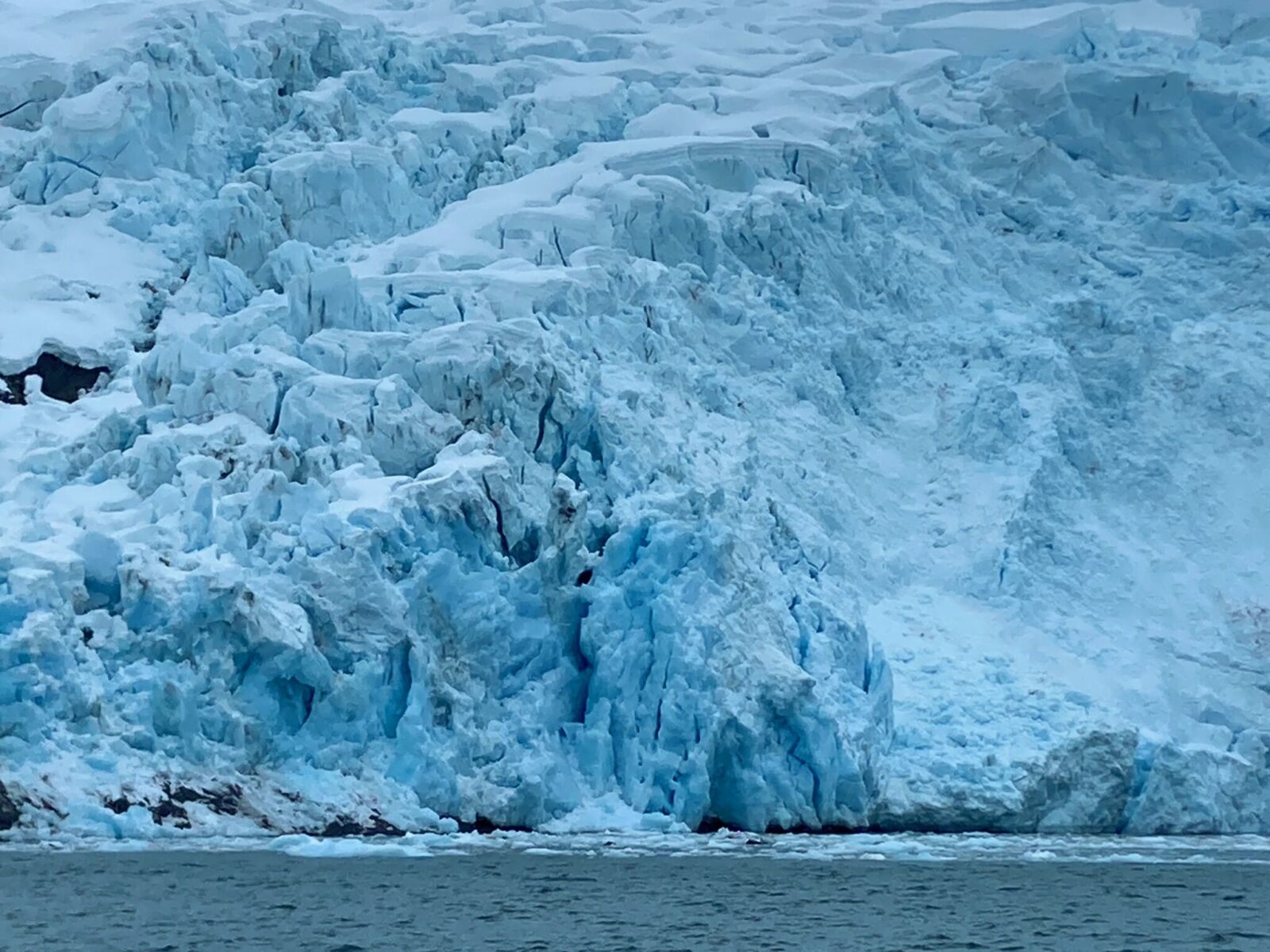 The crevases on the face of a tidewater glacier. They are white and blue and some dark rocks are poking out here and there. There are some icebergs in the water.