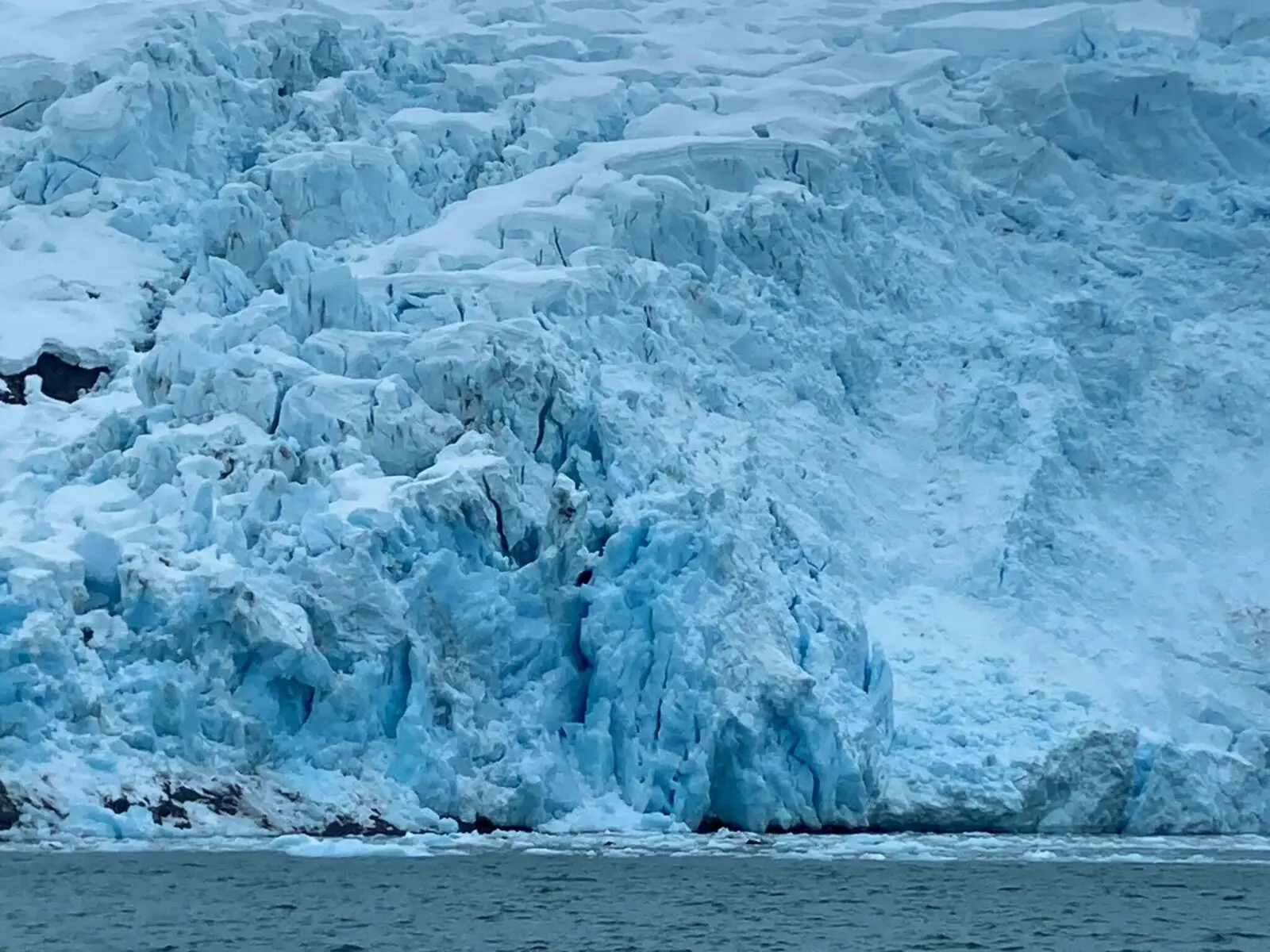 The crevases on the face of a tidewater glacier. They are white and blue and some dark rocks are poking out here and there. There are some icebergs in the water.