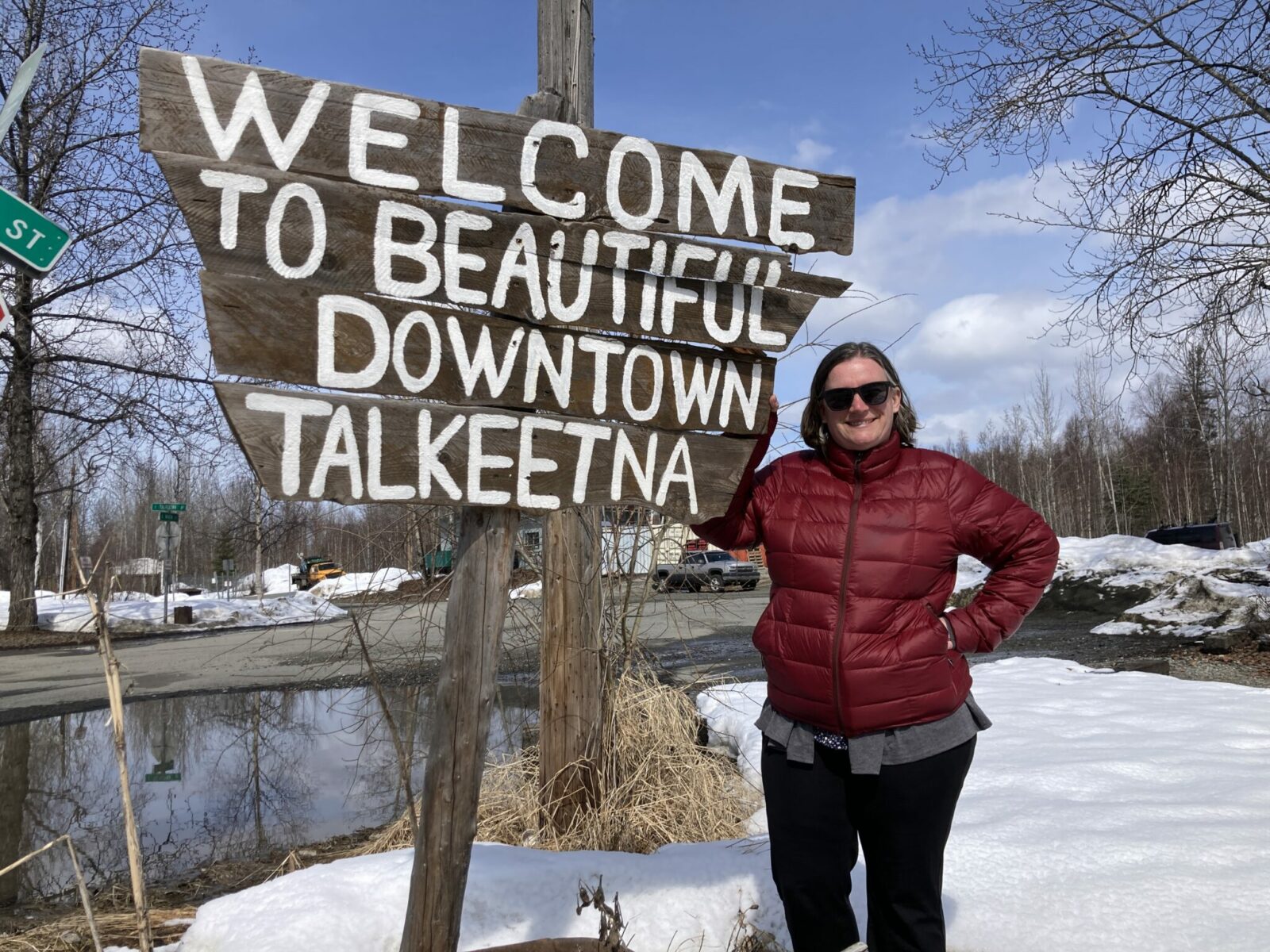 Jennie next to the sign that reads welcome to beautiful downtown Talkeetna. There is quite a bit of snow on the ground and the trees of not greened up yet