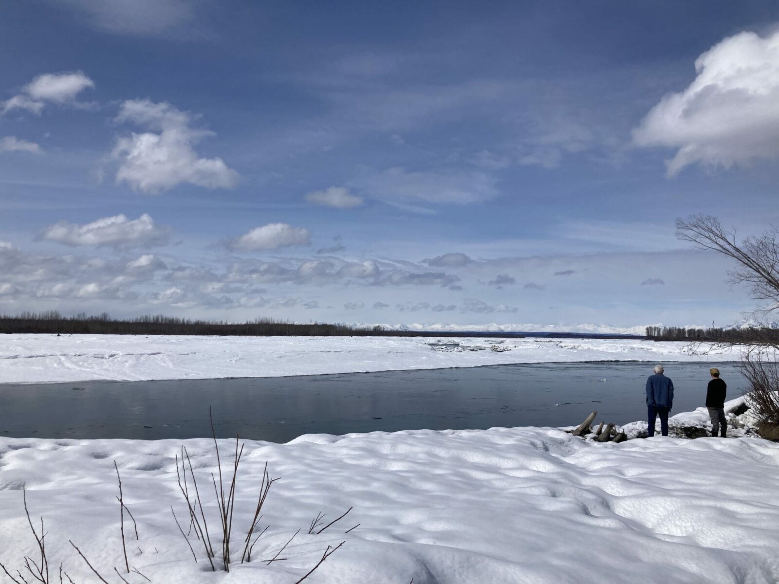 Snow banks in spring along the side of a wide river with mountains in the background. The river is flowing freely but still has some ice floating by.