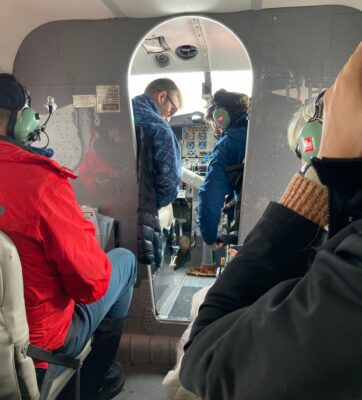 The interior of an Otter airplane, with several people sitting in seats with headsets. At the front you can see the pilot and a passenger in the cockpit buckling their seatbelts