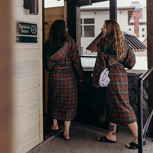 Two women in plaid robes with hoods and wearing slippers prepare to enter a sauna.