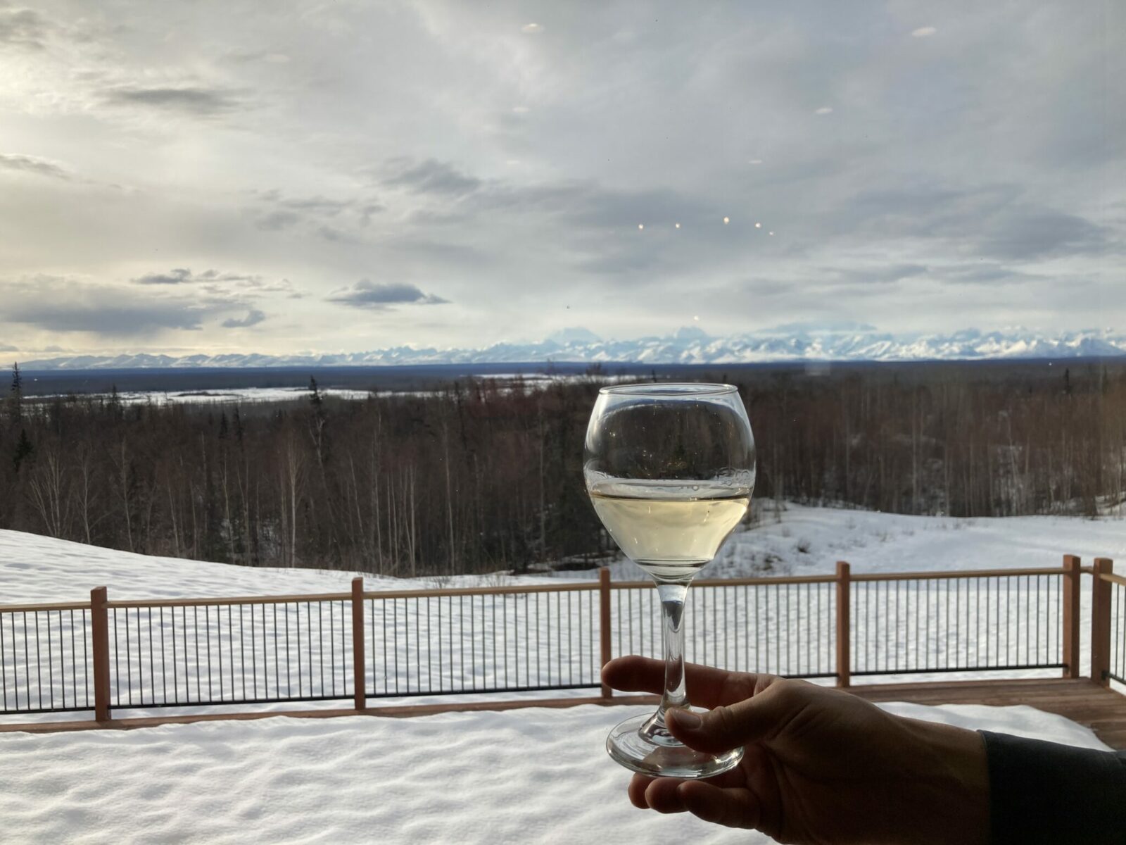 A glass of white wine being held up to the window with mountains and a deck covered in snow outside.