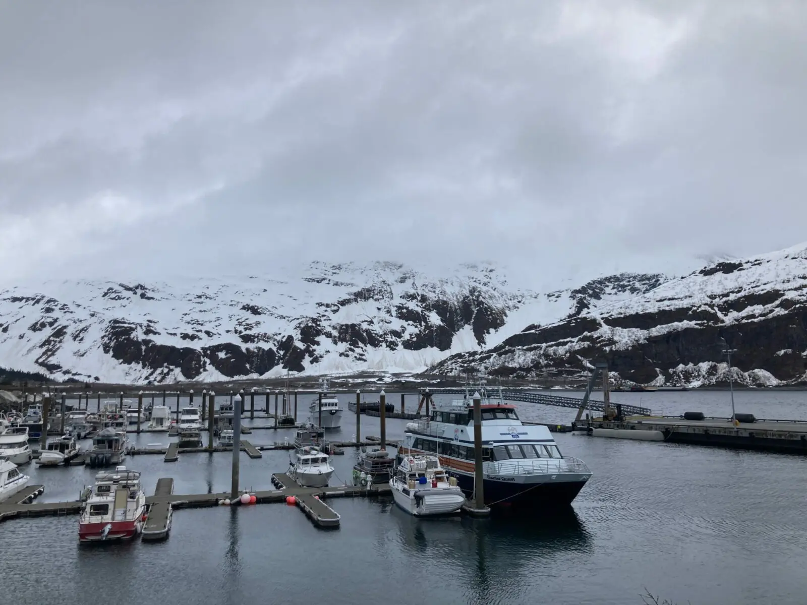 A small boat harbor with a few boats in it on an overcast day. There are snow covered hills around the harbor