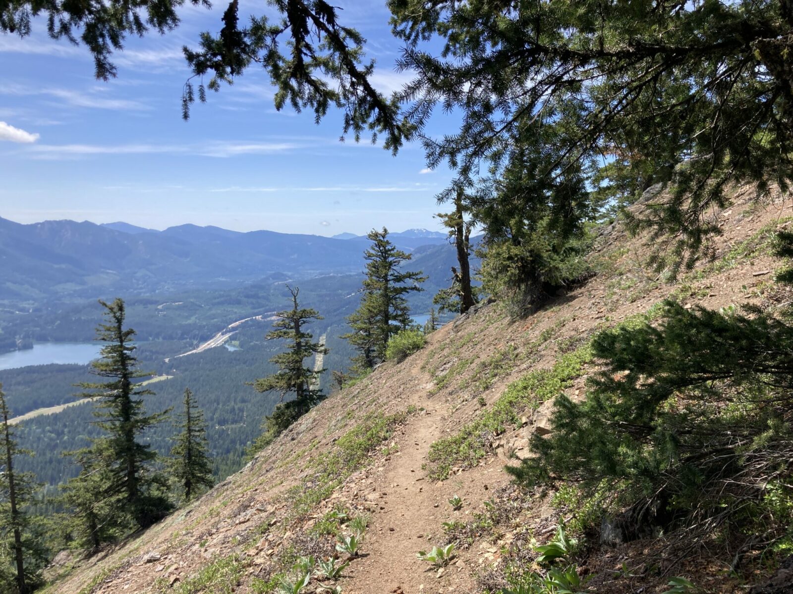 A narrow dirt trail cuts through a steep hillside with lakes and forest in the valley below along with a freeway