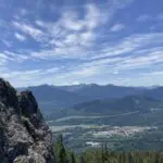 Mt Rainier is visible behind closer forested mountains and hills from the Easton Ridge Trail. The rocky summit of Easton Ridge is in the foreground and the town of Easton can be seen in the valley below.