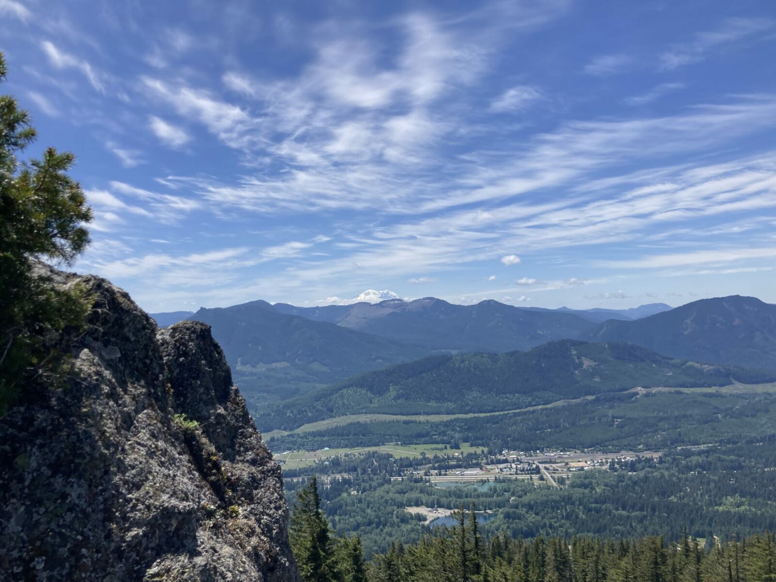 Mt Rainier is visible behind closer forested mountains and hills from the Easton Ridge Trail. The rocky summit of Easton Ridge is in the foreground and the town of Easton can be seen in the valley below.