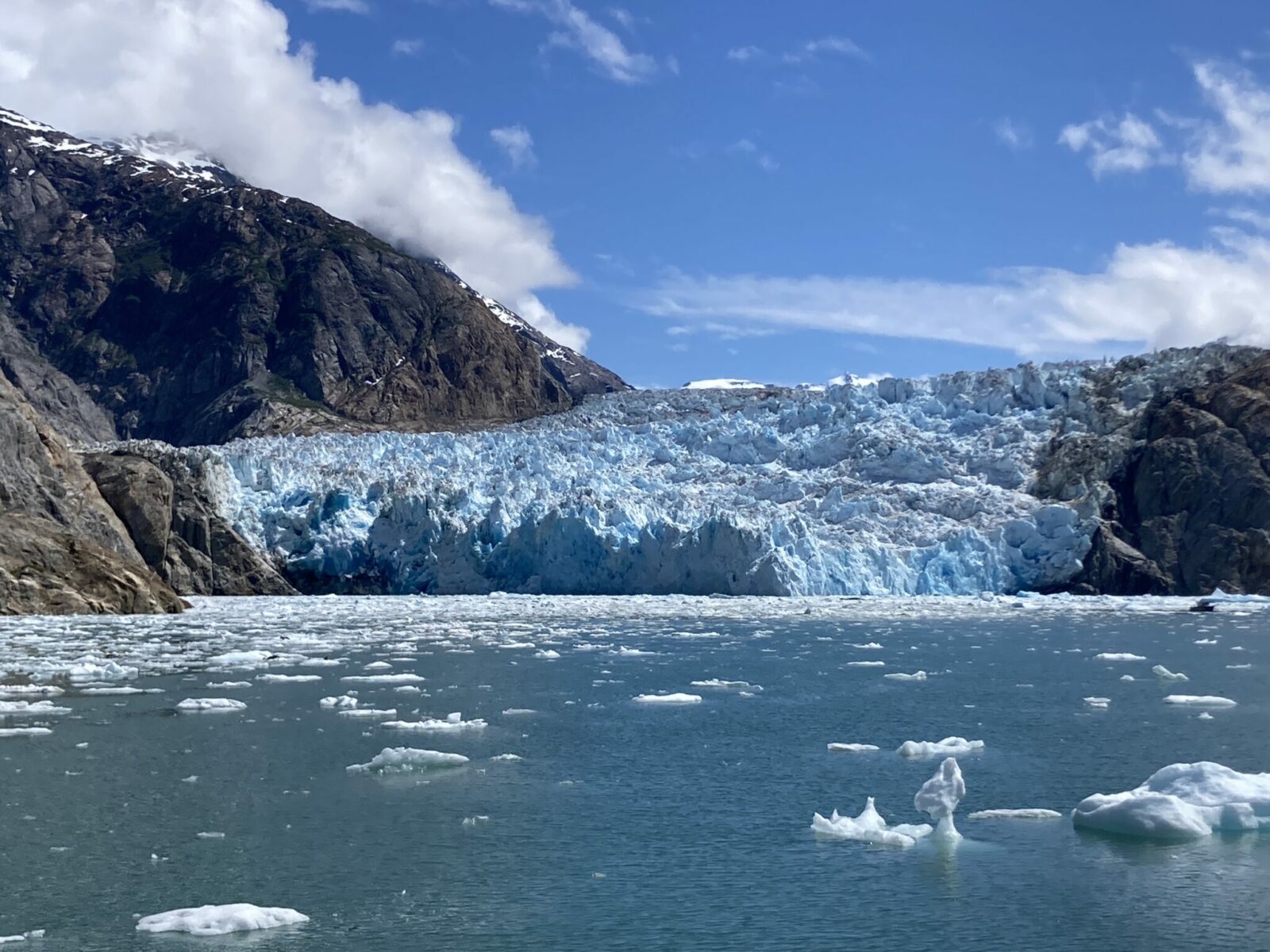 The blue ice of a tidewater glaciers at the head of a fjord surrounded by mountains on a sunny day.