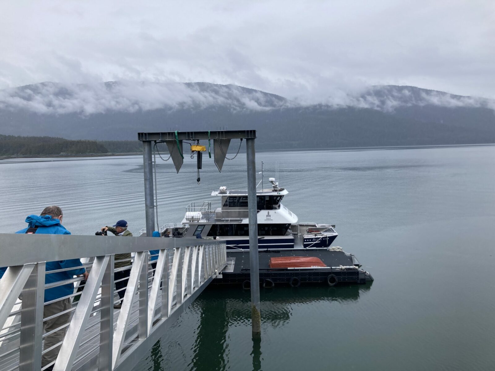 A white and dark blue tour boat docked at a floating dock with a few people walking down to the boat. There are forested hillsides in the background on a cloudy day