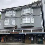 The exterior of a historic Victorian building, the Alaskan Hotel in Juneau. It is gray and has a bar downstairs and windows upstairs