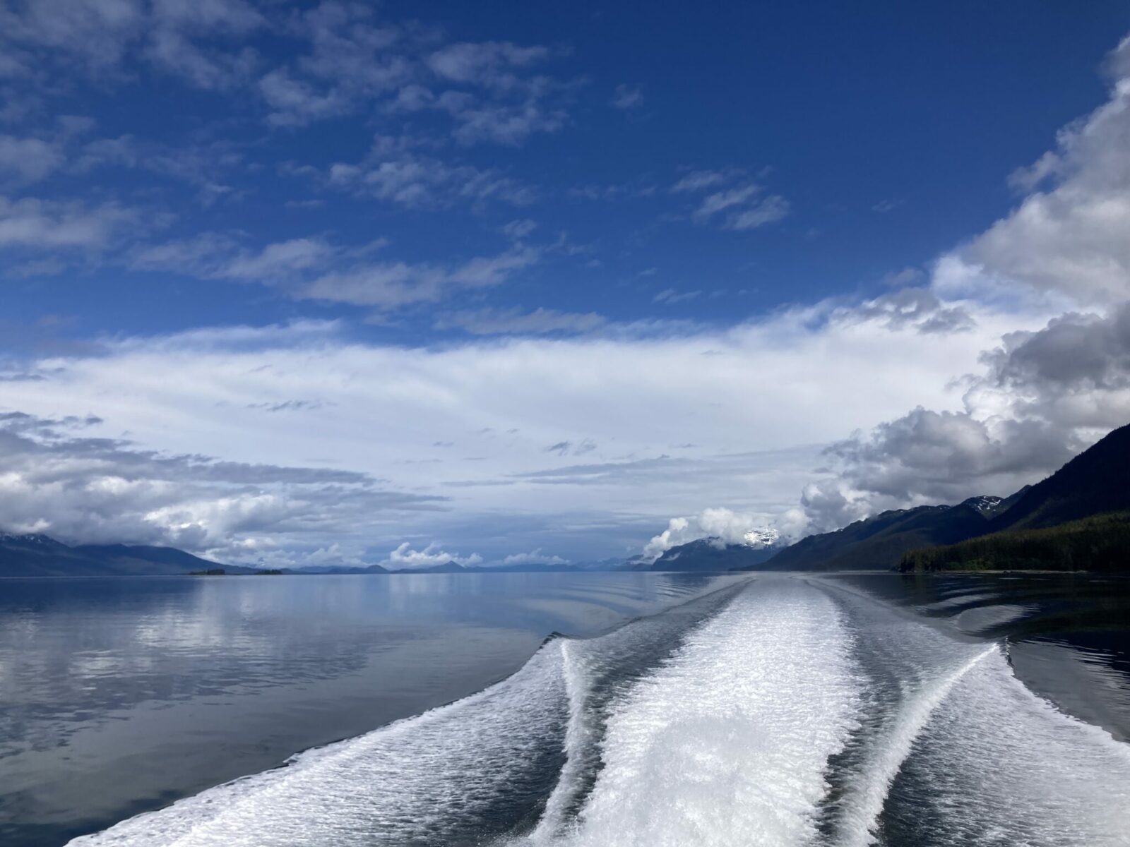 A photo from the back of a tour boat on the way back to Juneau from Tracy Arm. You can see the wake of the boat on calm water and mountains on both sides on a partly cloudy day