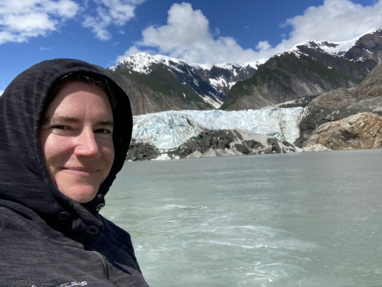 A person wearing a gray hooded fleece and smiling in front of a glacier and mountains. The water is milky from glacier silt.