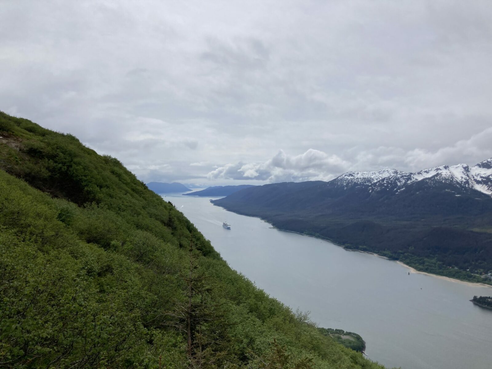 A view of a channel from a mountaintop with a cruise ship far below. Across the water are snow covered mountains. It is an overcast day