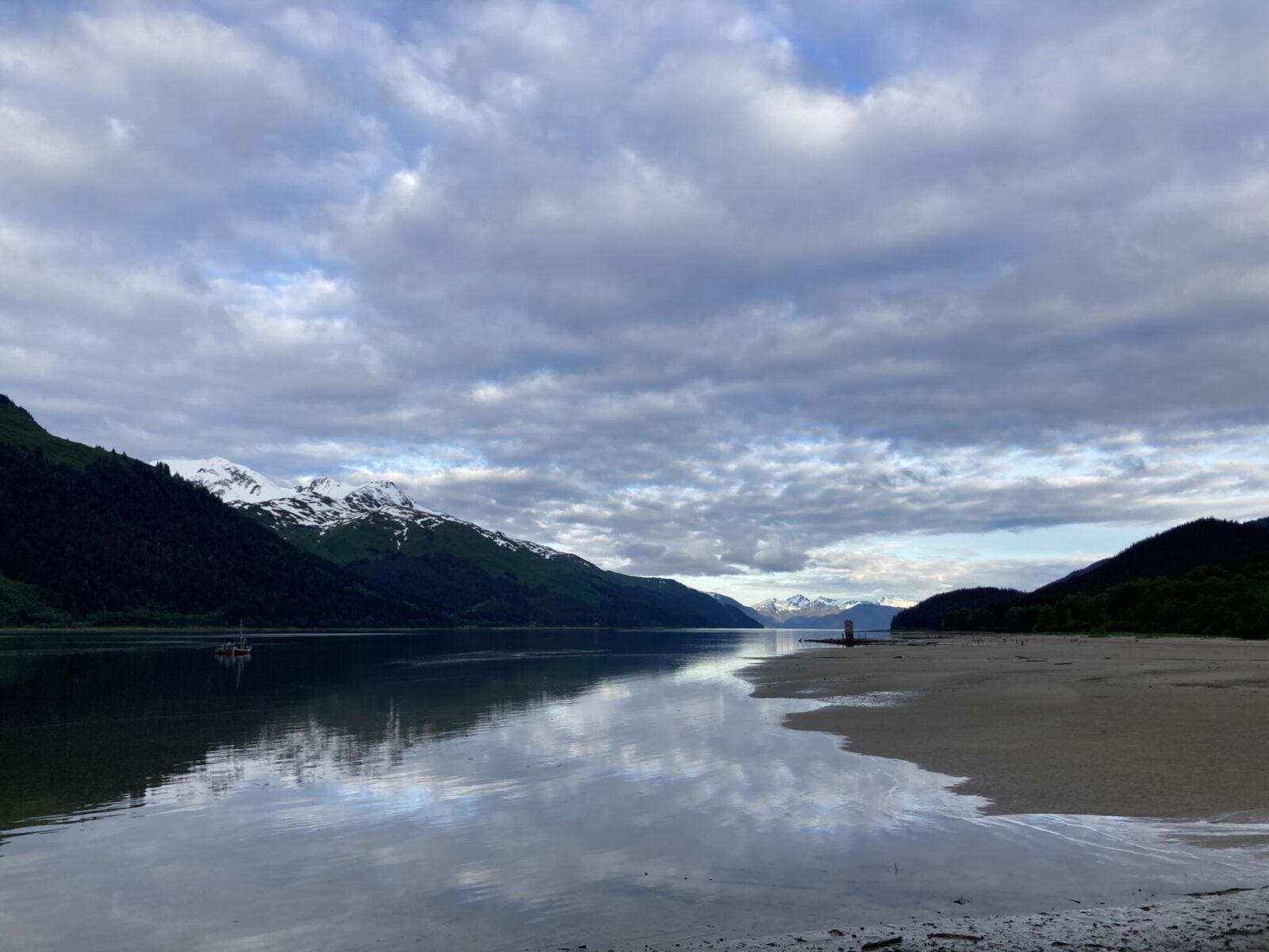 A sandy beach next to smooth water of a channel between two sets of mountains near Juneau Alaska at sunset on a mostly cloudy day