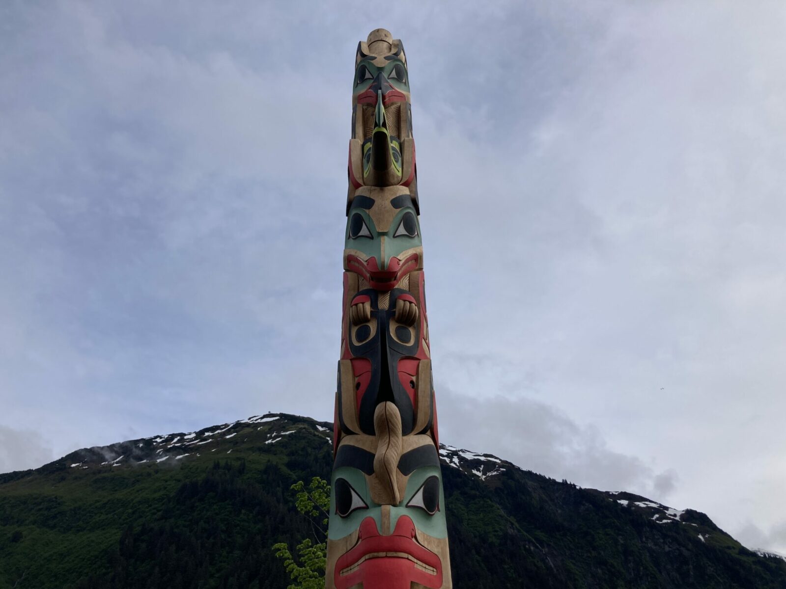 A totem pole with red, green and black figures with a mountain and some snow patches in the background in downtown Juneau Alaska