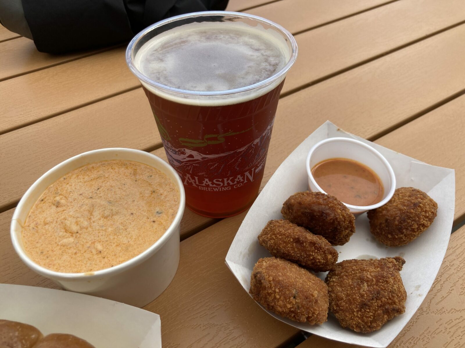 A cup of soup, five crab cakes and a beer on a picnic table