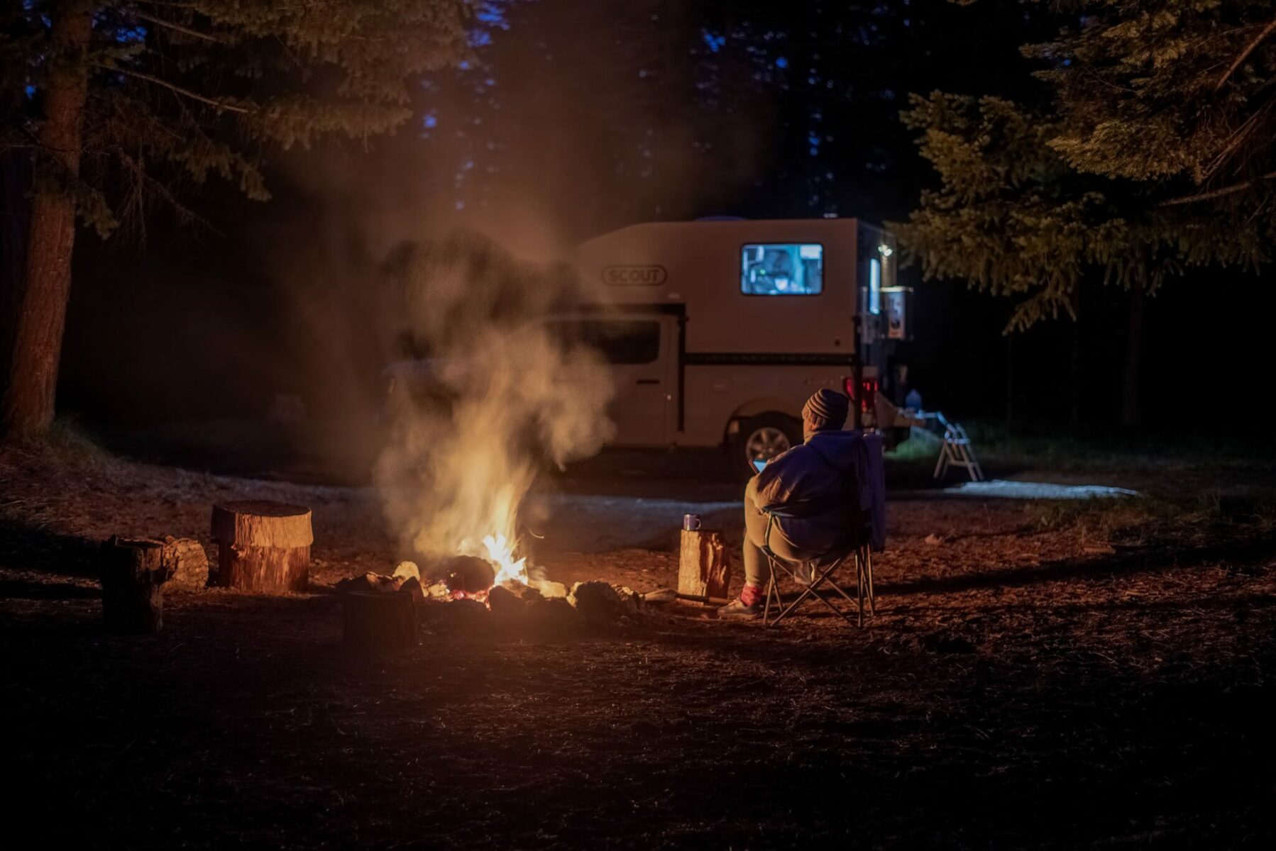 A person in a campchair sitting next to a campfire after dark with a pick up camper in the background