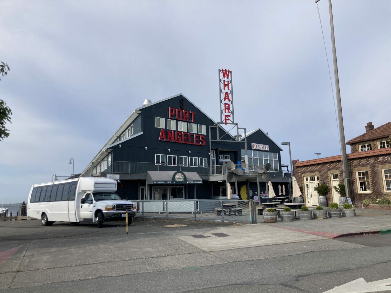 A blue building that says Port Angeles wharf with a brick building next door and a white airporter style bus parked in front.