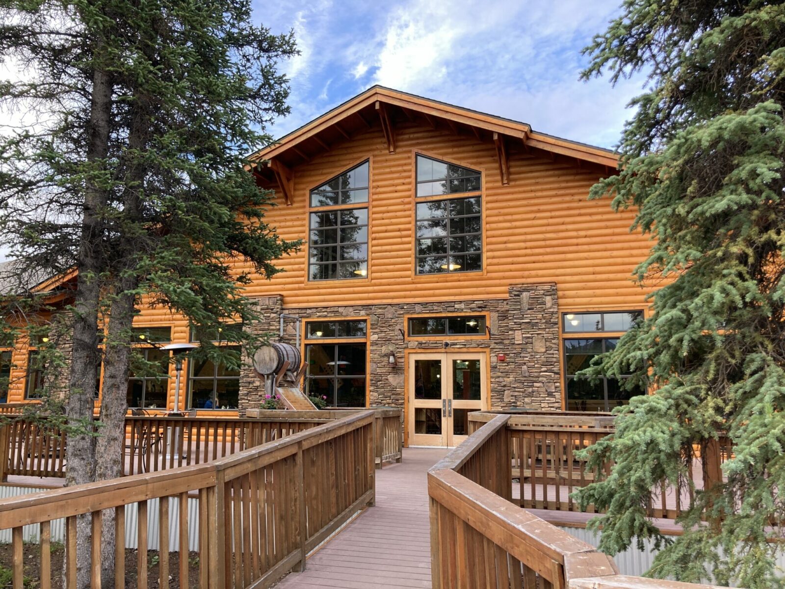 A wooden and stone lodge building with a wooden deck around it and a few trees