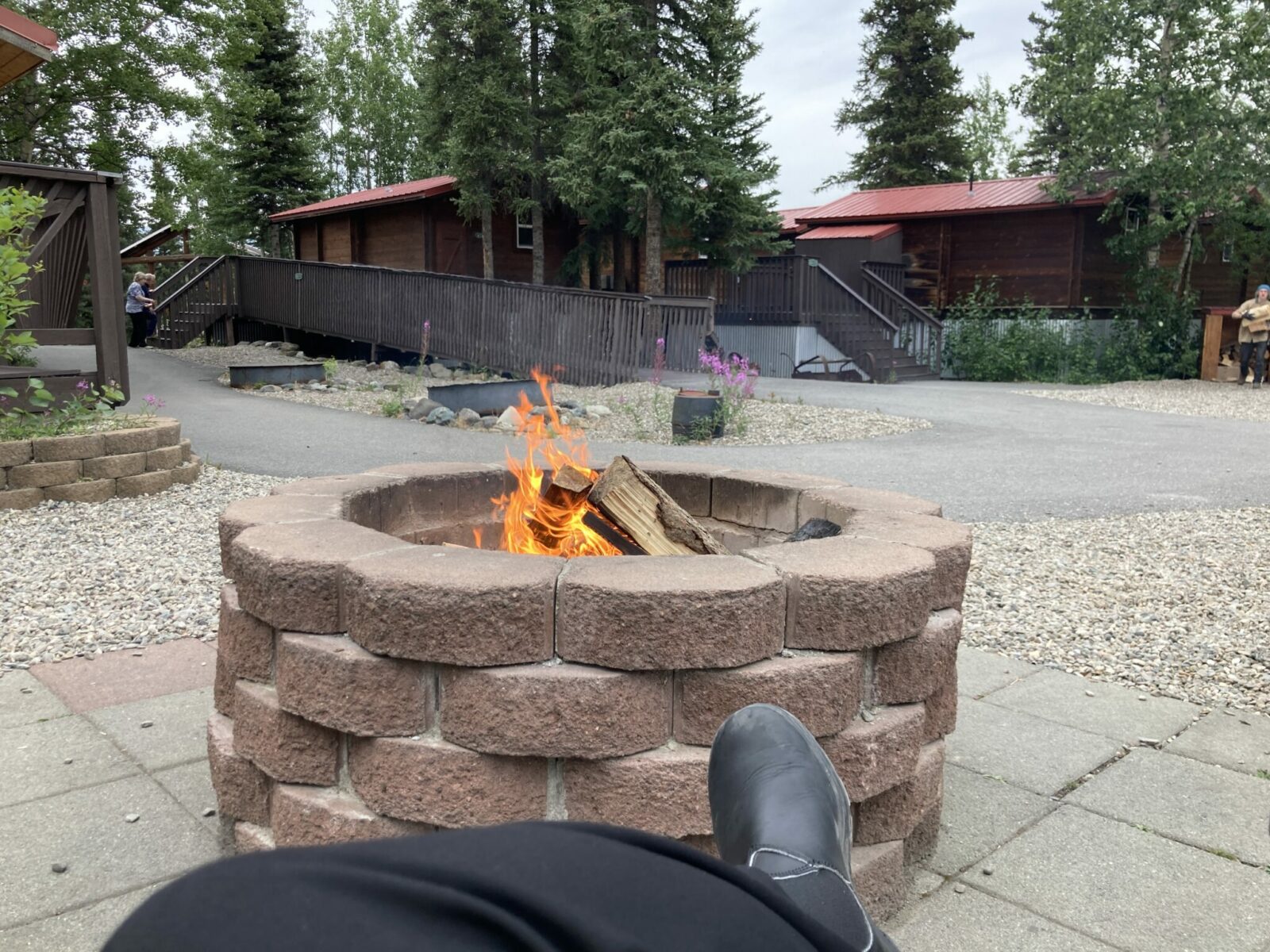 A stone fire pit with a fire in it and a person's feet sitting by it. Some cabins in the background