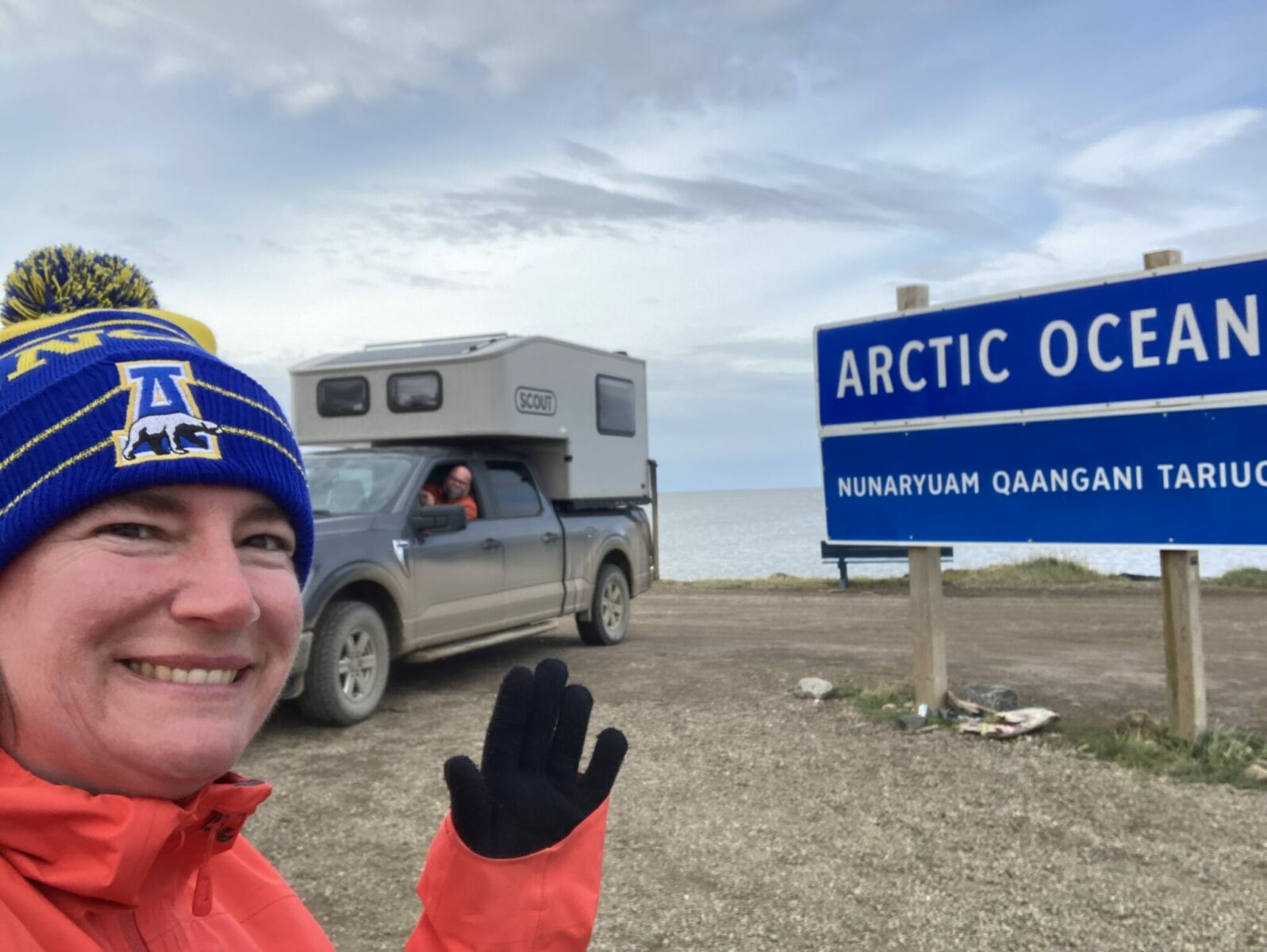 A person in a red rainjacket and blue hat smiles for the camera in front of a pick up camera at the beach next to the arctic ocean. A blue sign tells you its the arctic ocean