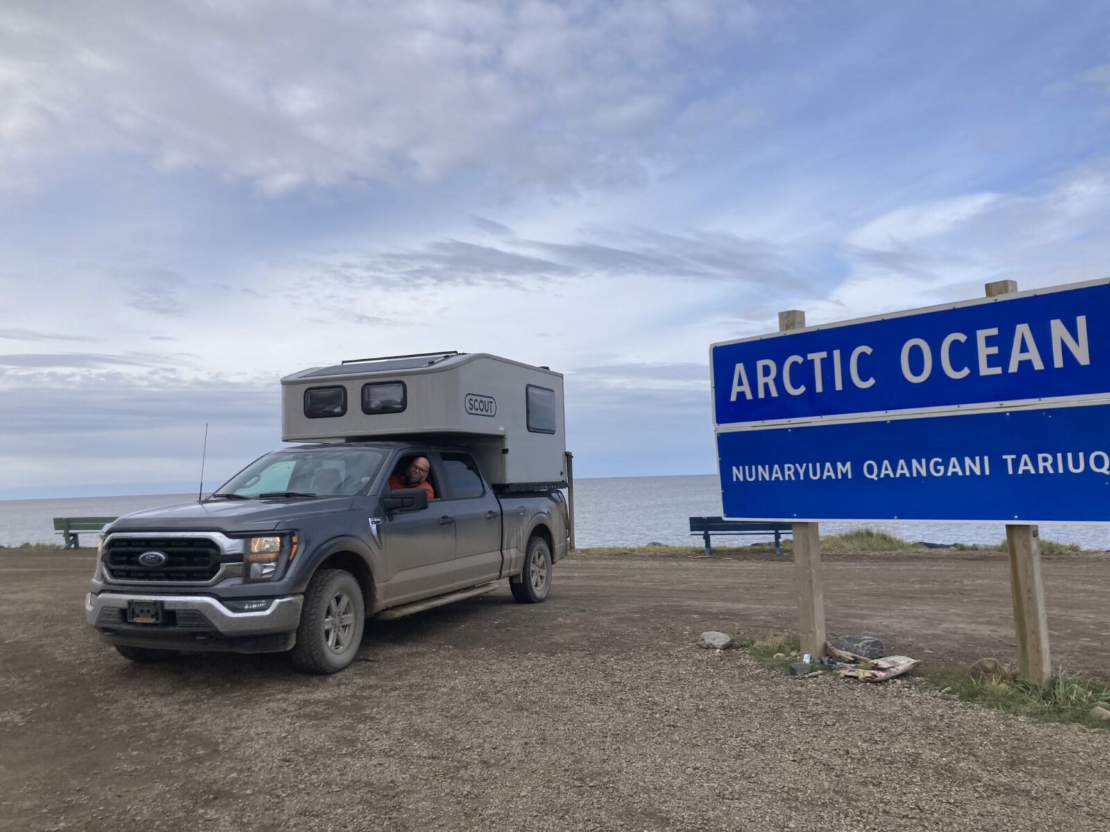 A person driving a gray pick up truck with a small camper in the bed parked on the beach next to a sign that says Arctic Ocean - Nunaryuam Qaangani Tariug