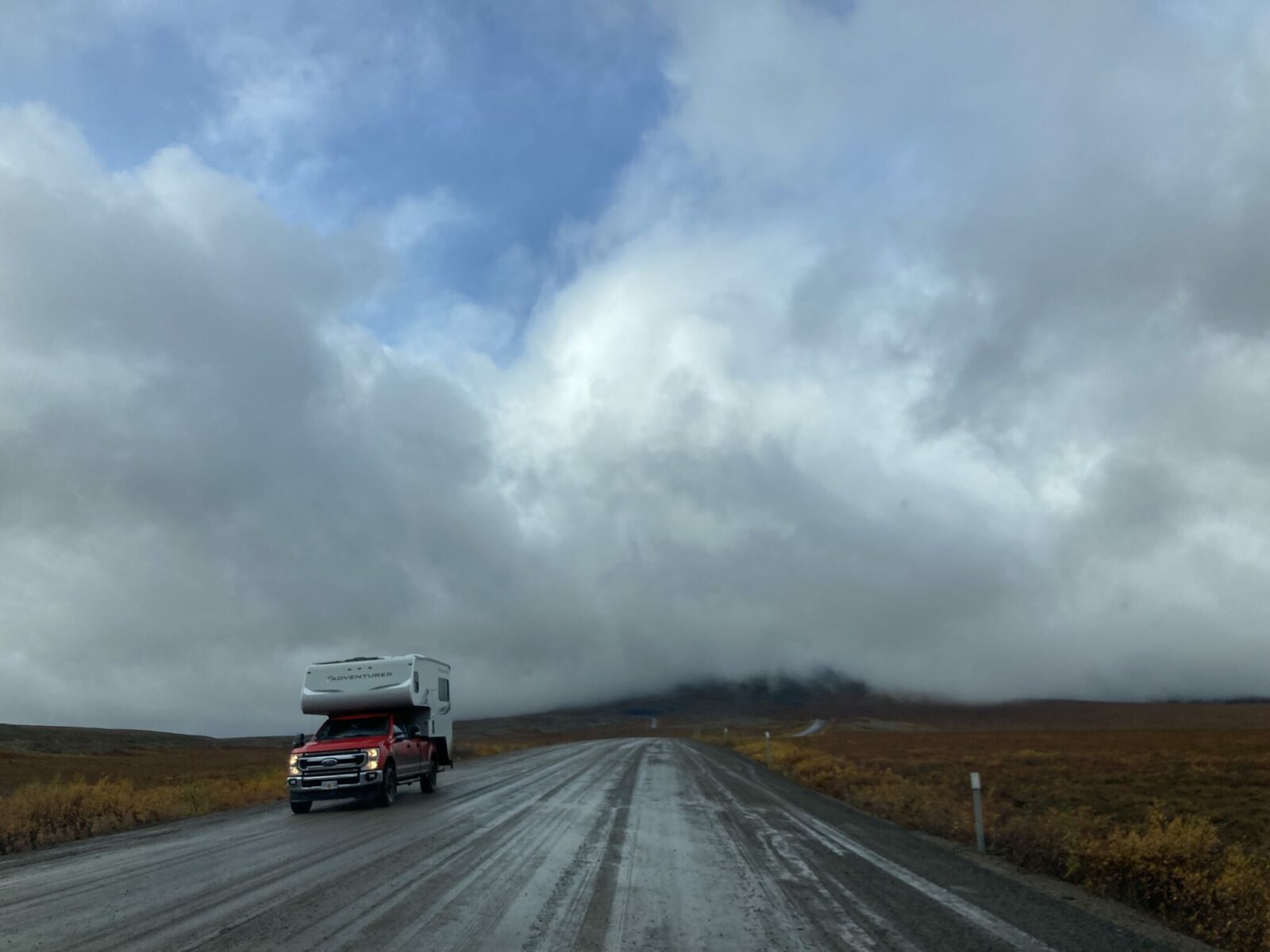 The Dempster Highway with fog rolling in. The road is muddy and wet and there is a pick up camper driving. on it.