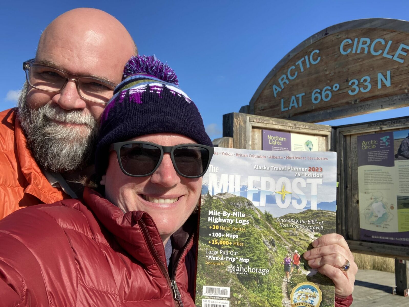 A close up selfie of two people holding up a Milepost in front of the Arctic Circle sign on the Dempster Highway