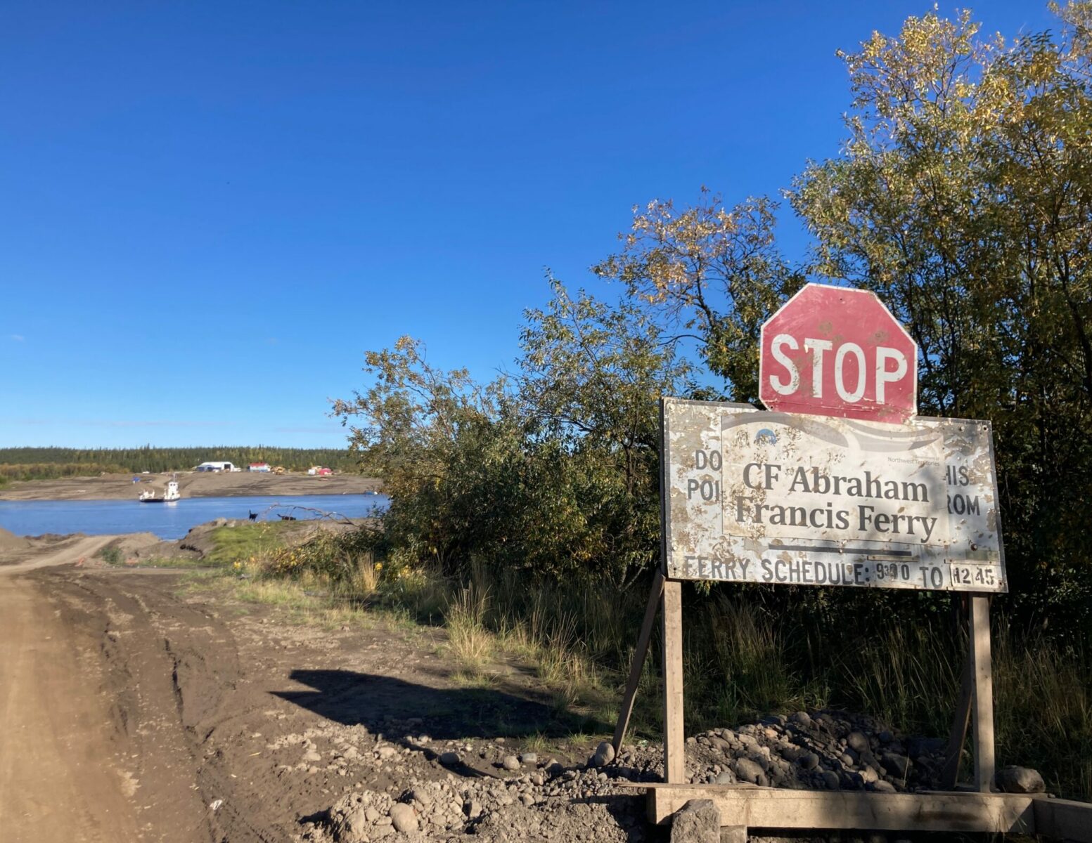 A stop sign at a ferry on a gravel ramp at the Peel River on the Dempster highway. You can see a few buildings and a cable ferry in the background.